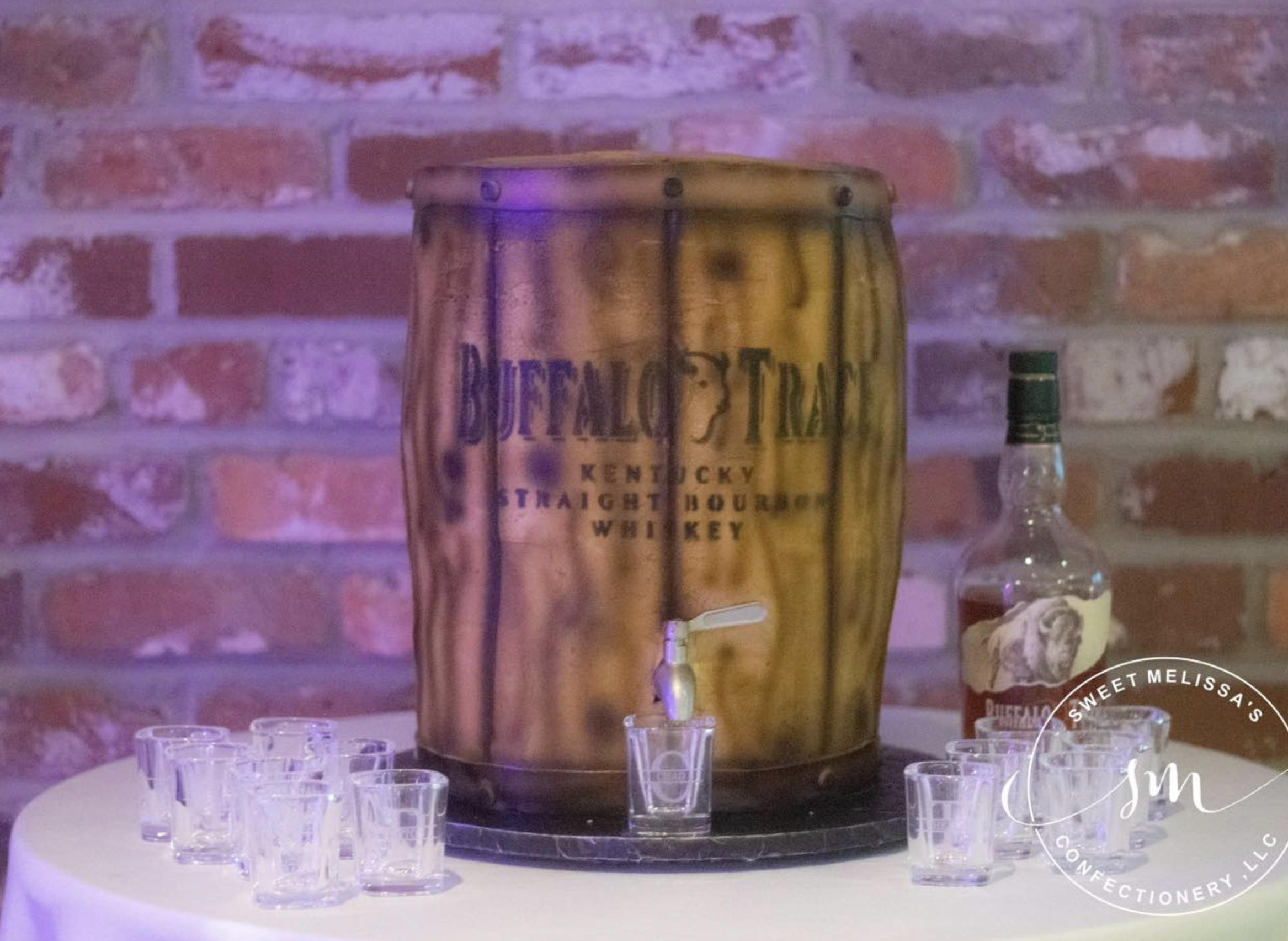 A round table set up with a large metal cooler labeled 'Old Forester Kentucky Straight Bourbon Whiskey' in the center and several small shot glasses surrounding it. In the background, there is a brick wall and a bottle of liquor on the right side. Th