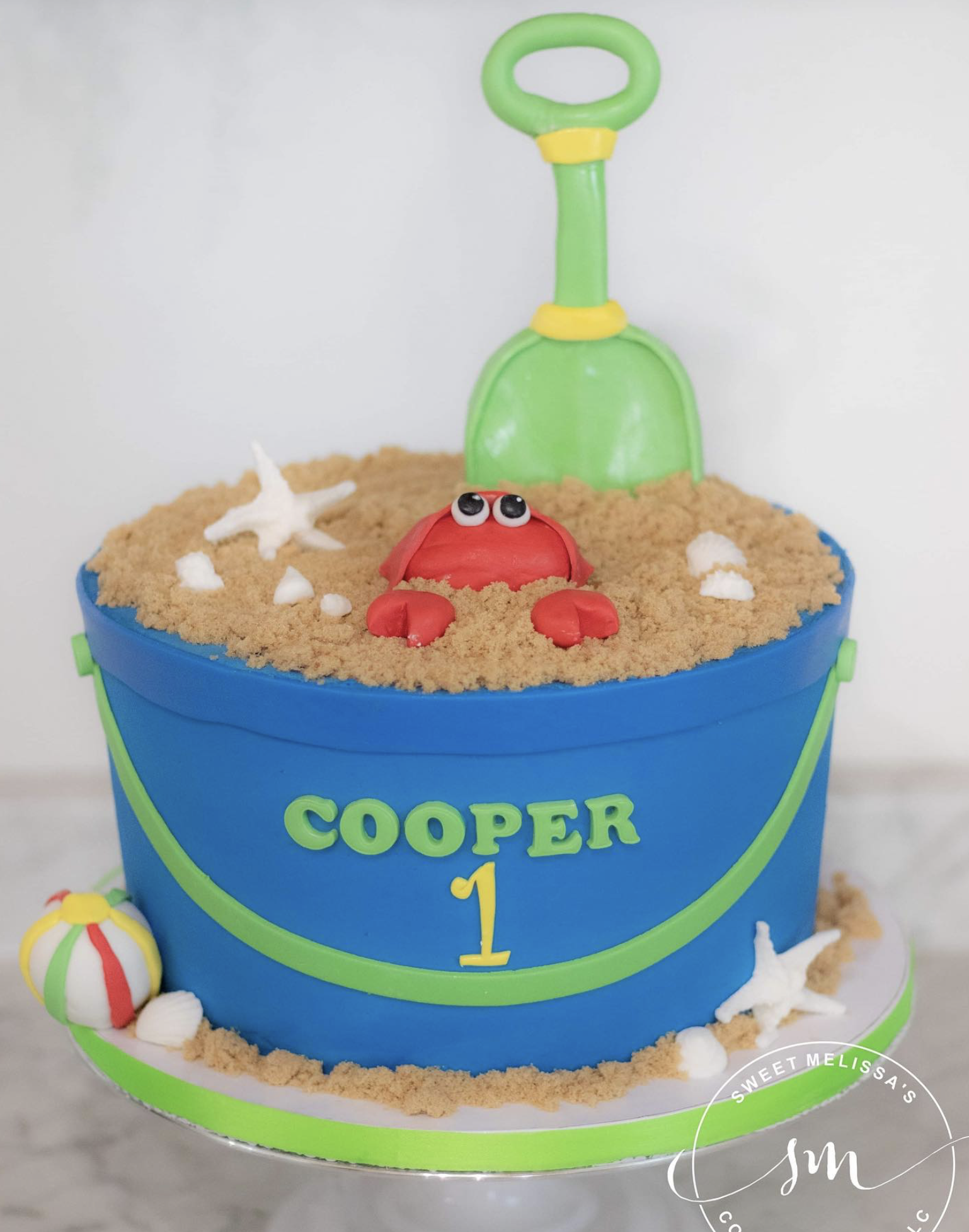 A birthday cake decorated with a beach theme, featuring a red crab, a white starfish, a striped beach ball, and a yellow and white beach umbrella, with a blue bucket labeled 'Cooper 1' as the main part of the cake.