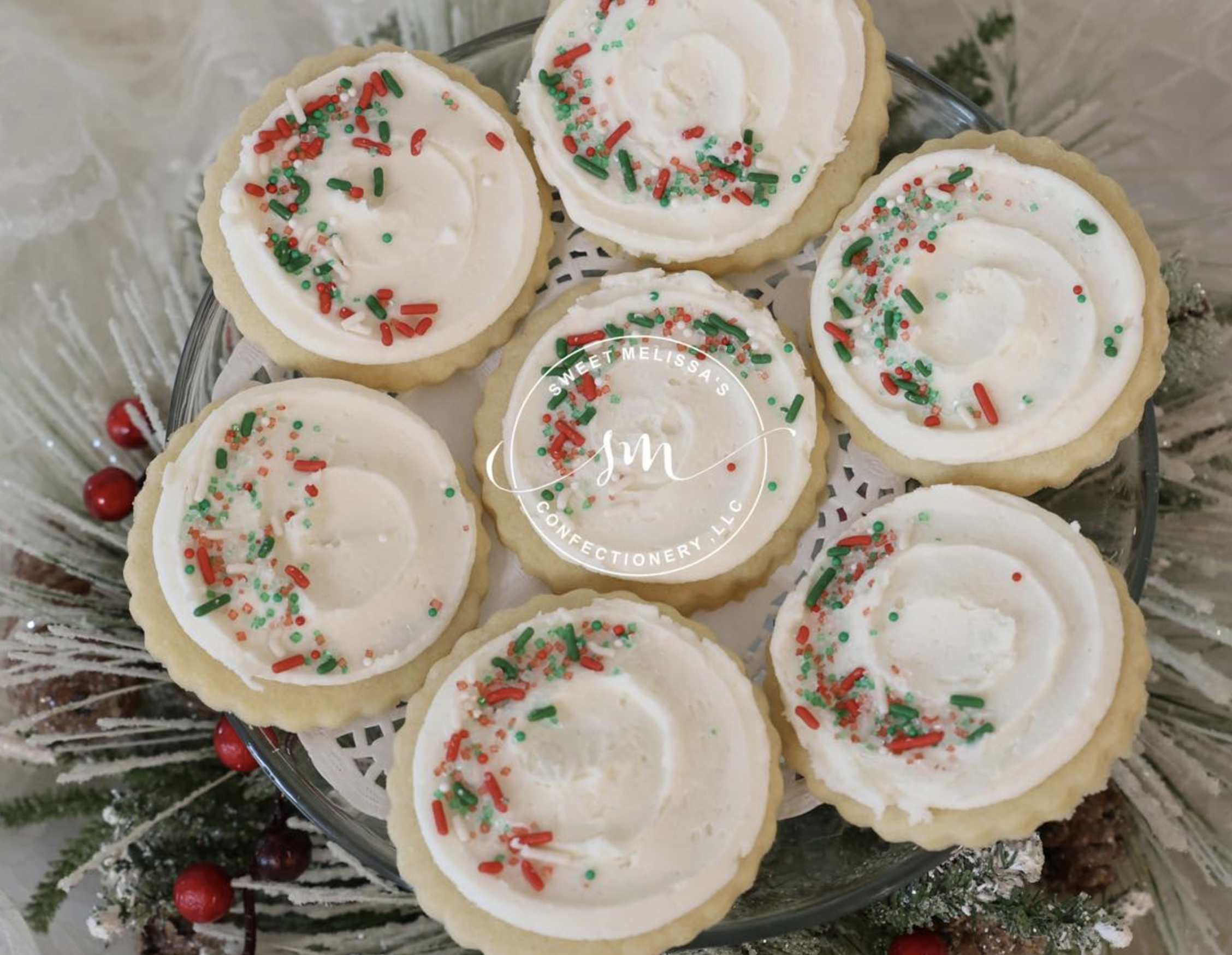 A glass platter of sugar cookies topped with white frosting and colorful sprinkles, surrounded by Christmas decorations including red berries and evergreen branches.