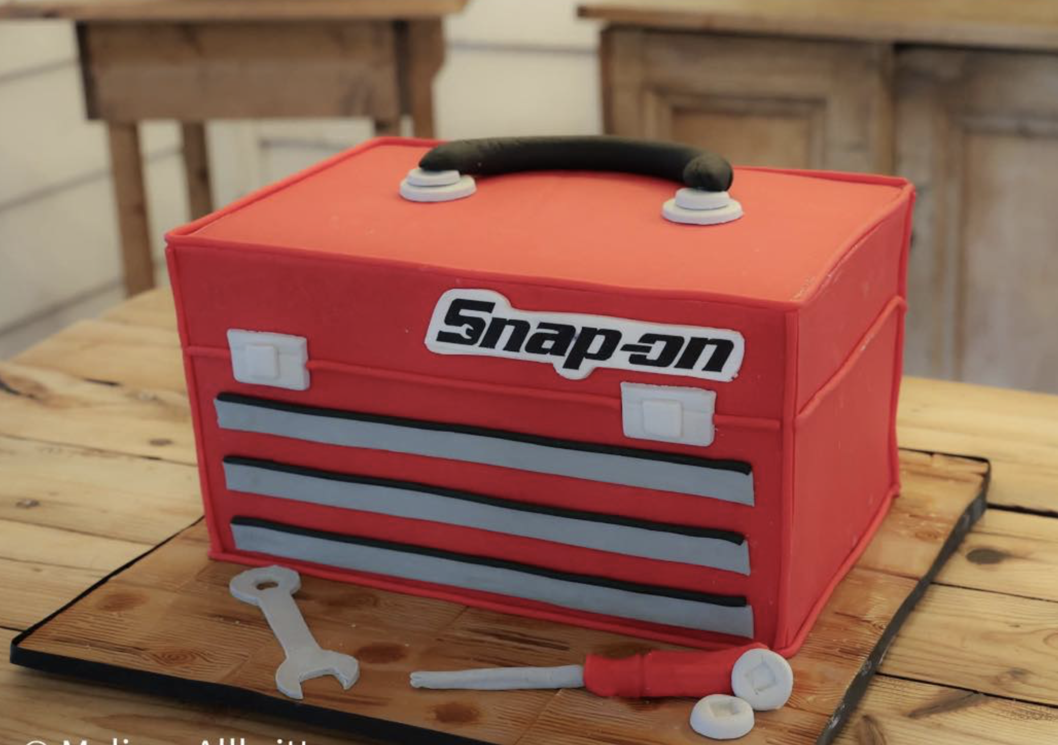 A red tool box with the Snap-on logo on the front, placed on a wooden table. There are white plastic wheels and a white plastic wrench in front of the toolbox, and a small red bottle behind it.