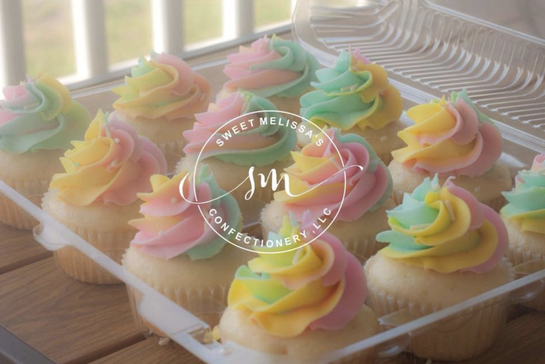 A clear plastic container of colorful cupcakes with pastel swirl frosting in pink, yellow, and green, on a wooden surface with a window in the background.