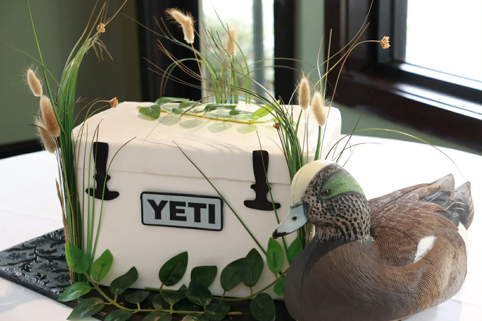A white cake with the YETI logo, decorated with greenery and tall grass, topped with a duck figurine, and placed on a black tray with a black and white pattern.