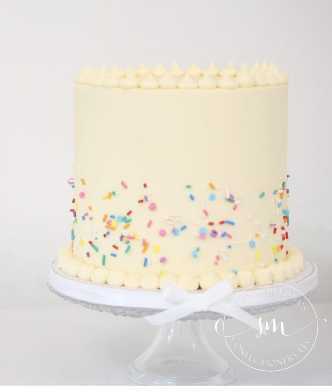 A tall, white, cylindrical birthday cake decorated with colorful sprinkles and piped cream on top, displayed on a glass cake stand.