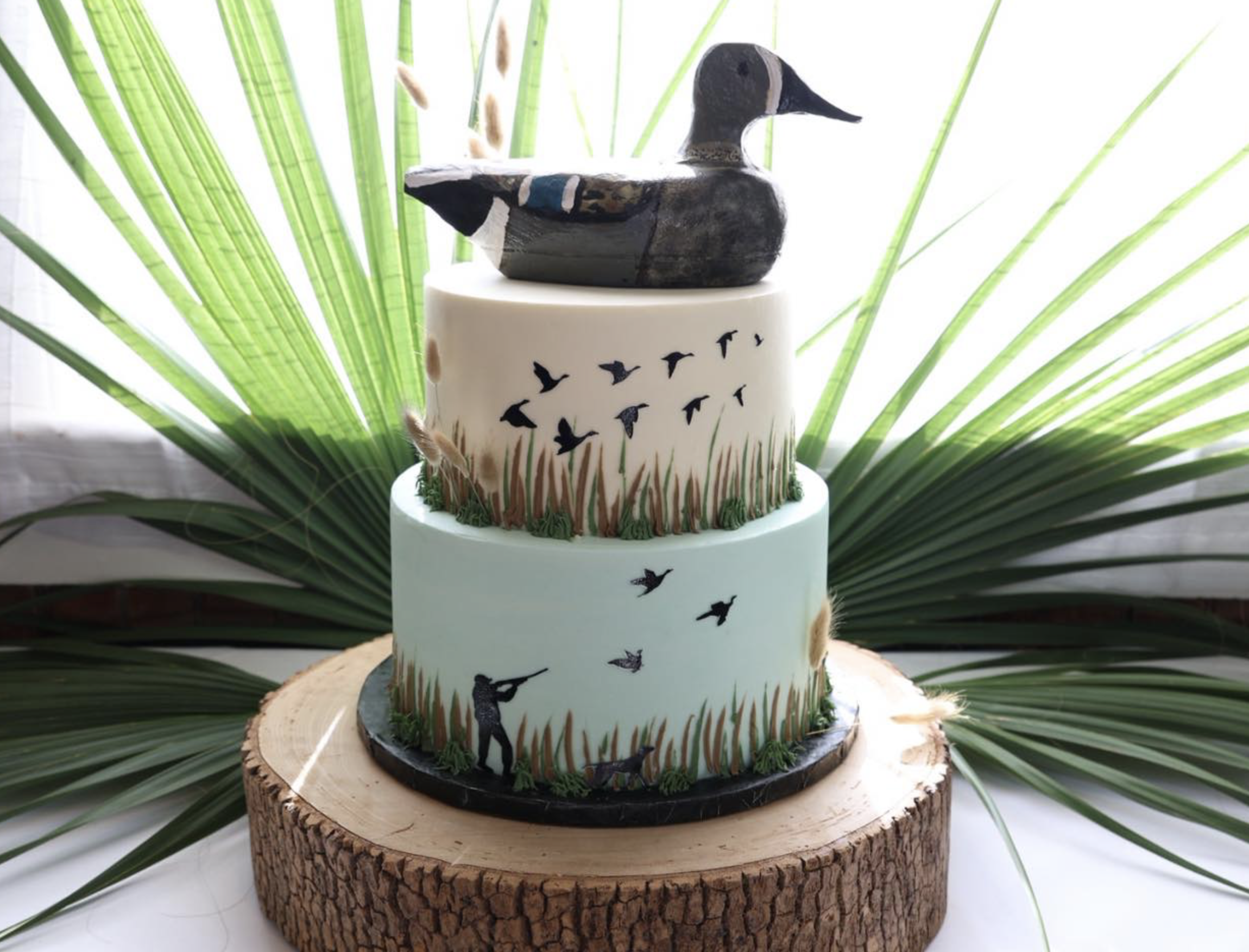 A multi-tiered cake decorated with a duck figurine on top, surrounded by a forest scene, set on a wooden log base with green palm leaves in the background.