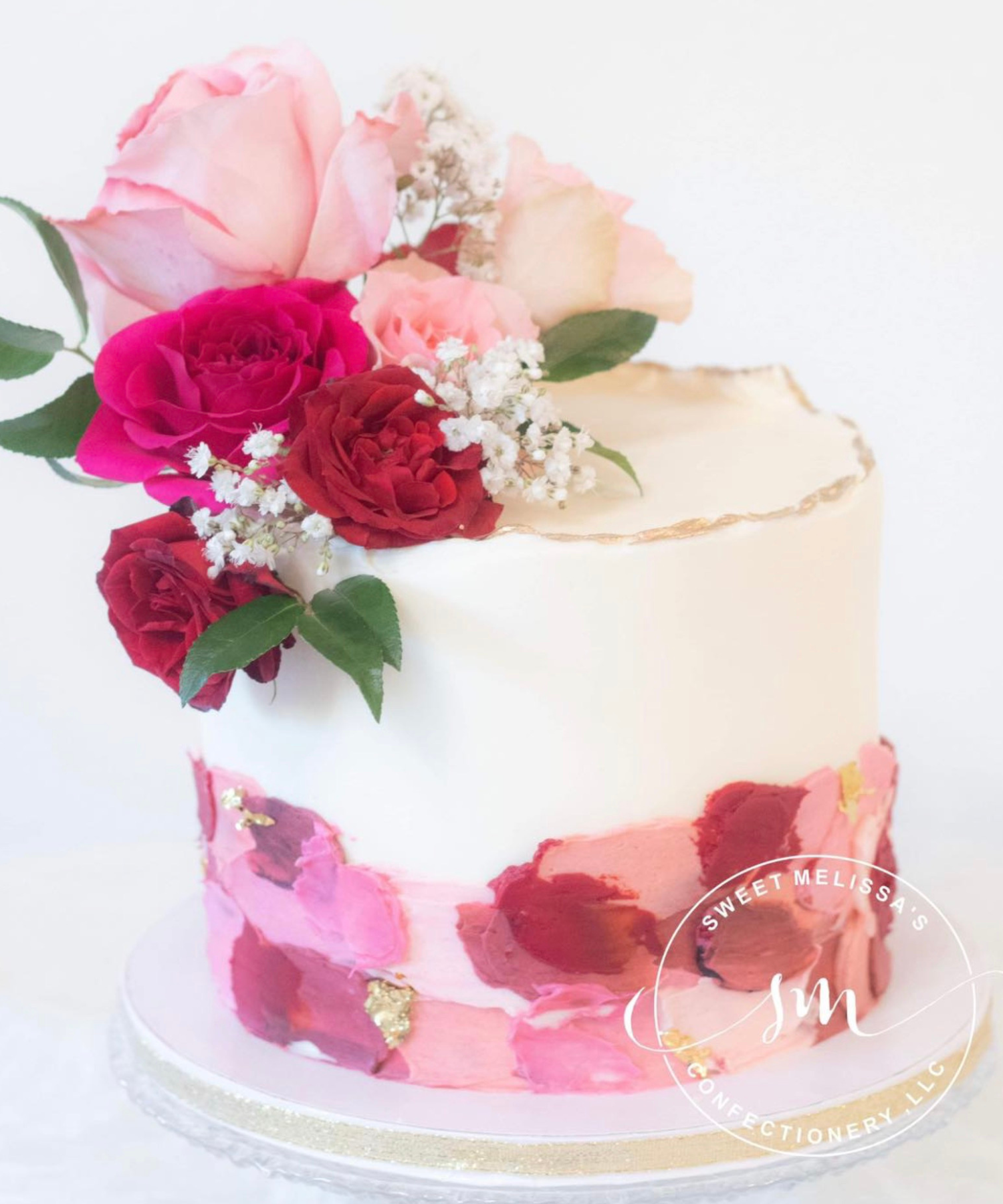 A pink and white decorated cake topped with a bouquet of pink, red, and cream roses, white baby's breath, and green leaves. The cake has artistic pink and red frosting decorations and gold accents.
