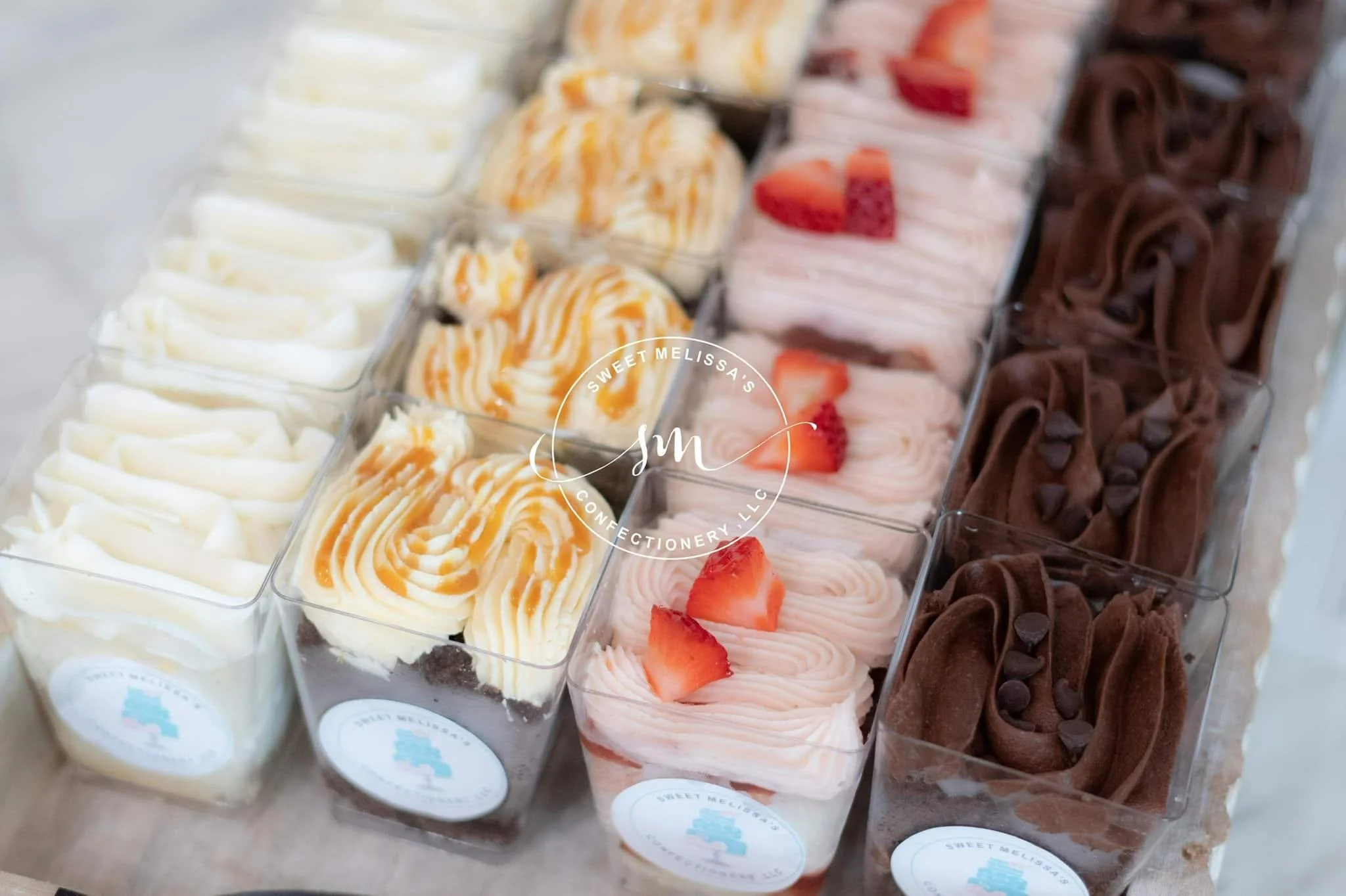 Assorted flavors of piped mousse or frosting in clear plastic containers, decorated with small toppings, arranged in a white box with a pink rose and a red rose nearby.