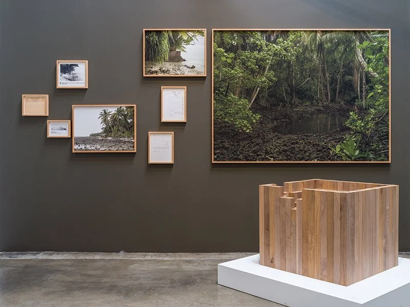 Gallery wall with framed photographs and texts depicting natural landscapes, with a wooden geometric sculpture in front.