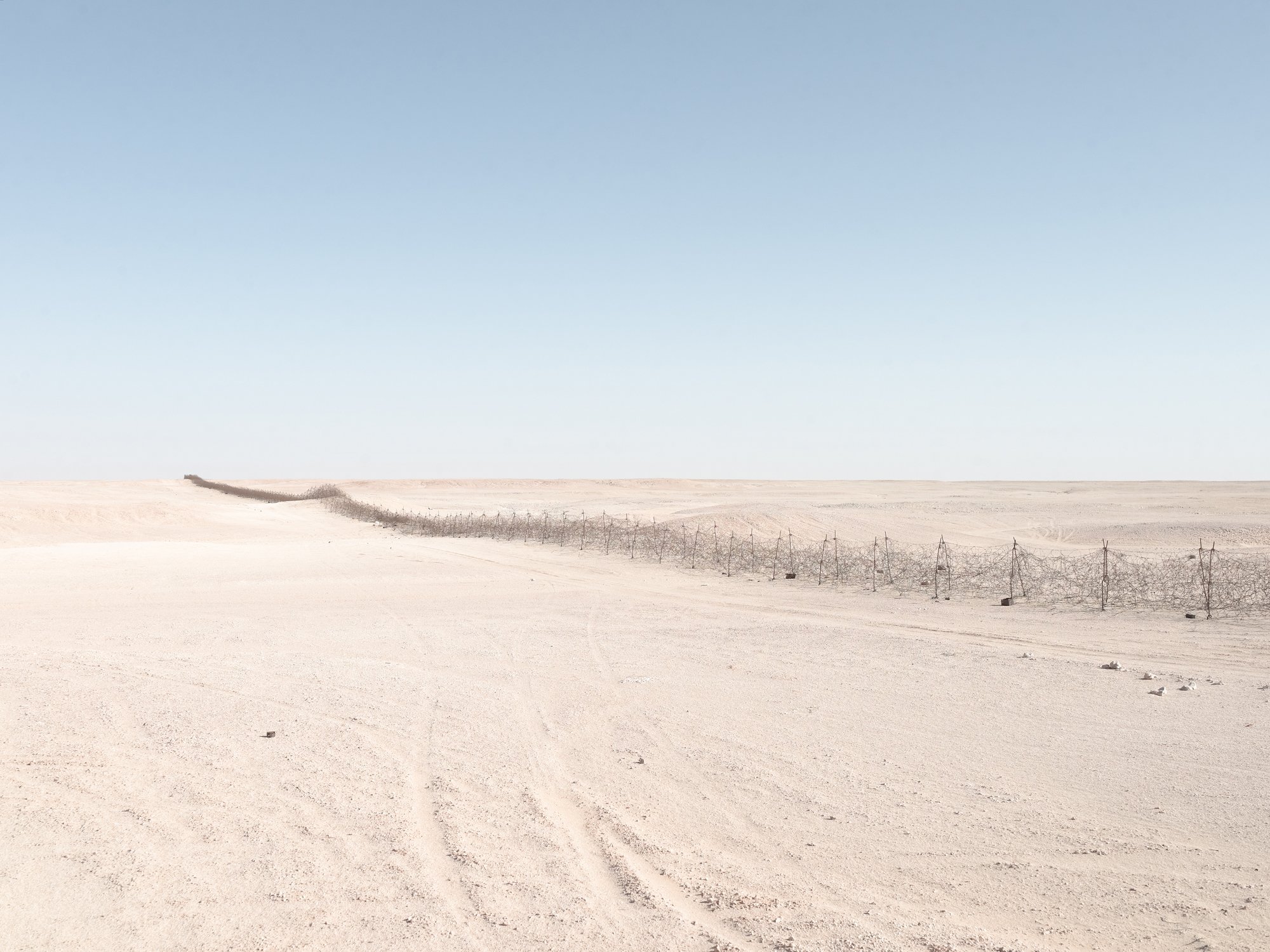 A vast, flat desert landscape with white sands and a clear blue sky, featuring a dirt road and a wire fence running across the scene.