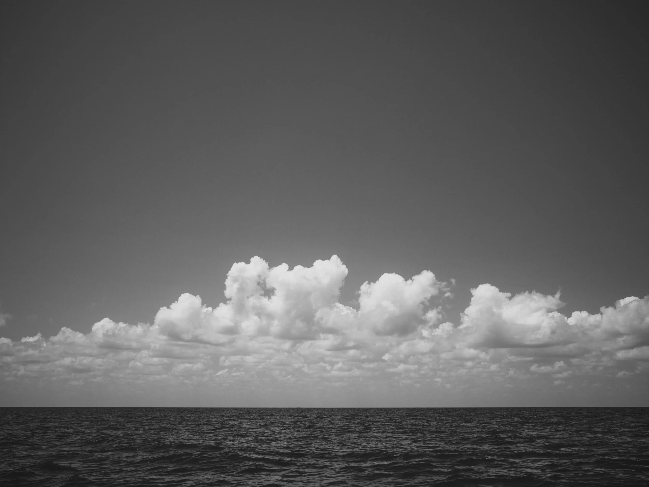Black and white image of a seascape with a horizon line, a sky filled with clouds, and a large body of water in the foreground.