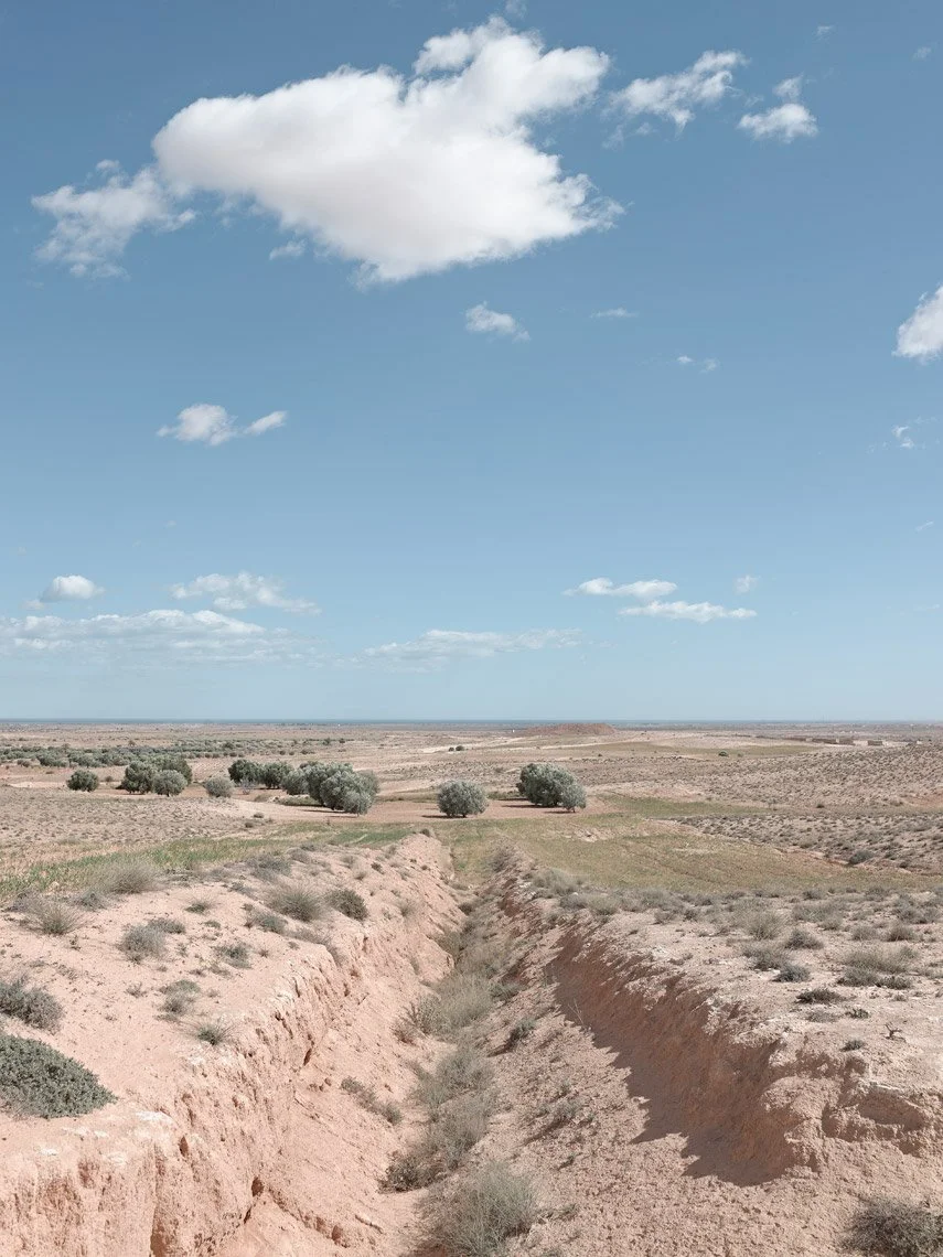 A dry, arid landscape with sparse vegetation and a small green patch, under a blue sky with scattered white clouds.