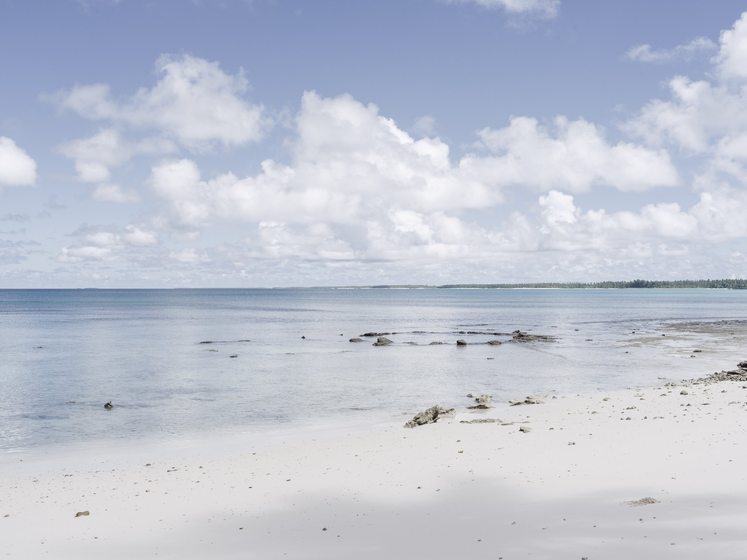 A beach scene with white sand, scattered rocks, and calm ocean water under a partly cloudy sky.