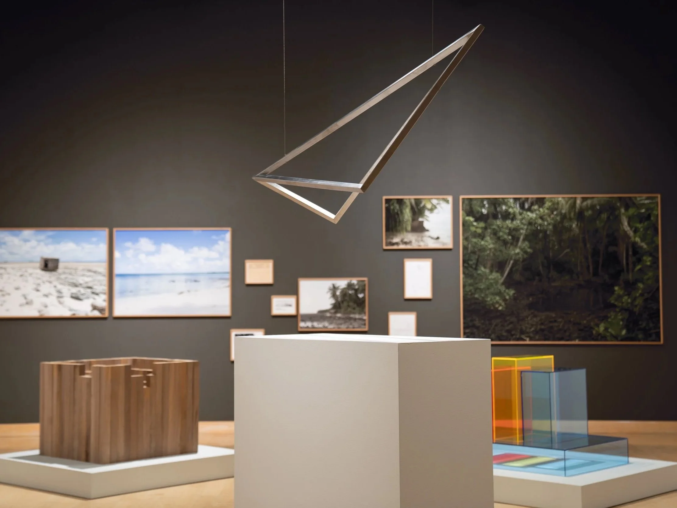 Artwork installation with framed nature photographs on dark wall, geometric wooden and glass sculptures, and colorful translucent boxes on white pedestal.