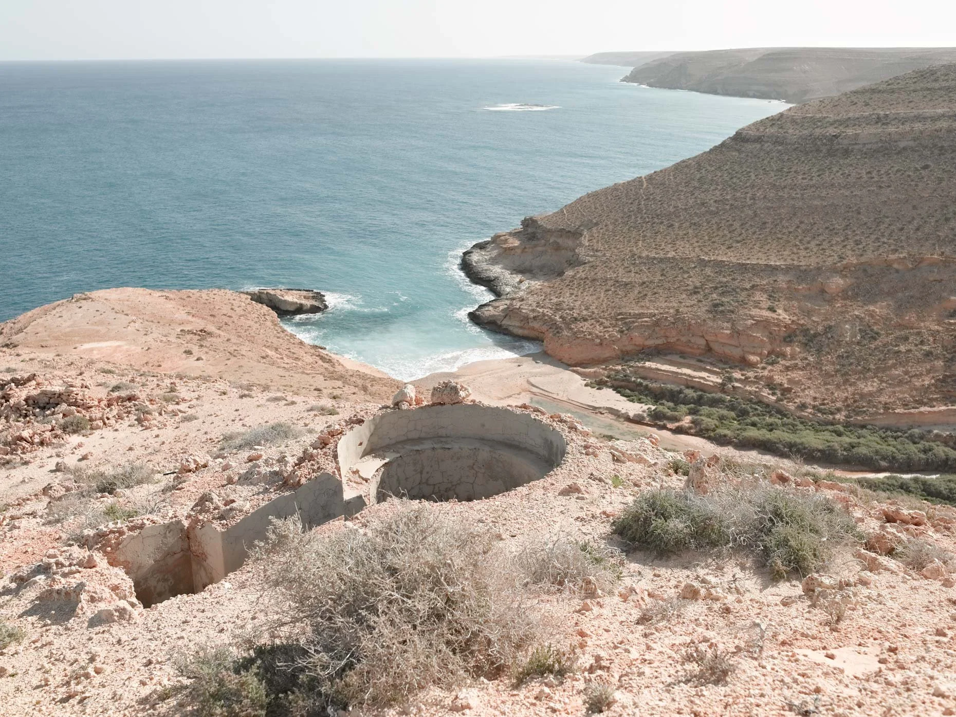 A desert coastline with rugged cliffs and hills overlooking the ocean. An old, abandoned well or cistern is in the foreground with arid, sparse vegetation surrounding it.