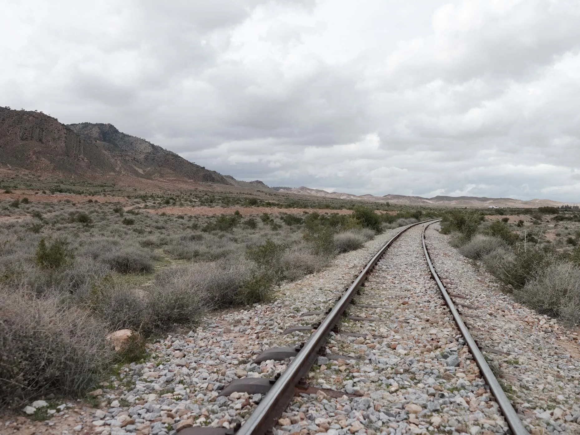A railway track running through a dry, barren landscape with low bushes and mountains in the distance under a cloudy sky.