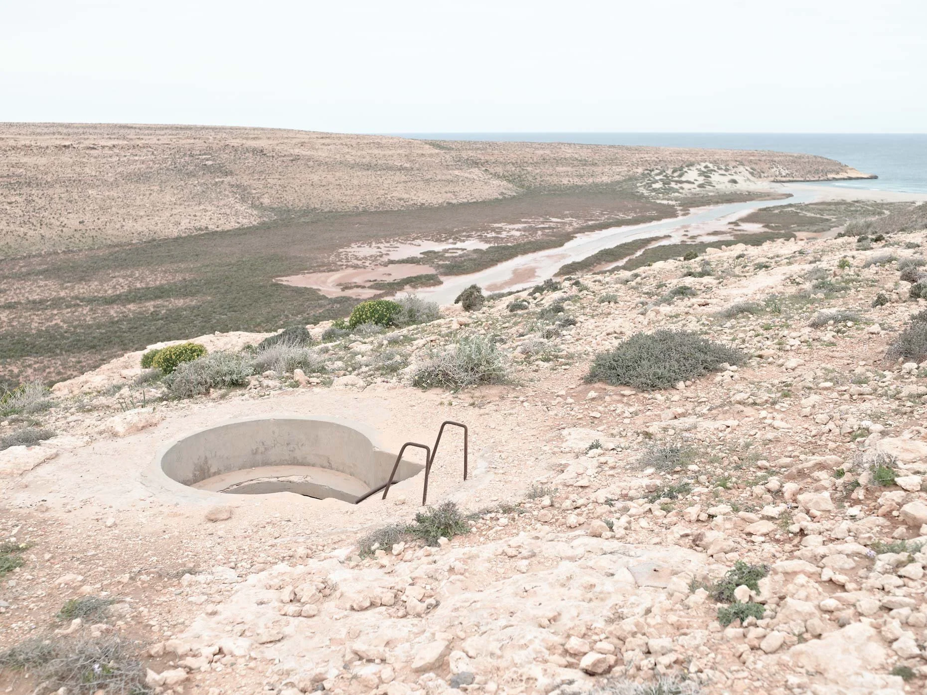 A desert landscape with sparse vegetation, a circular entrance to a bunker in the foreground, and a view of hills leading to a coastline with the ocean in the background.