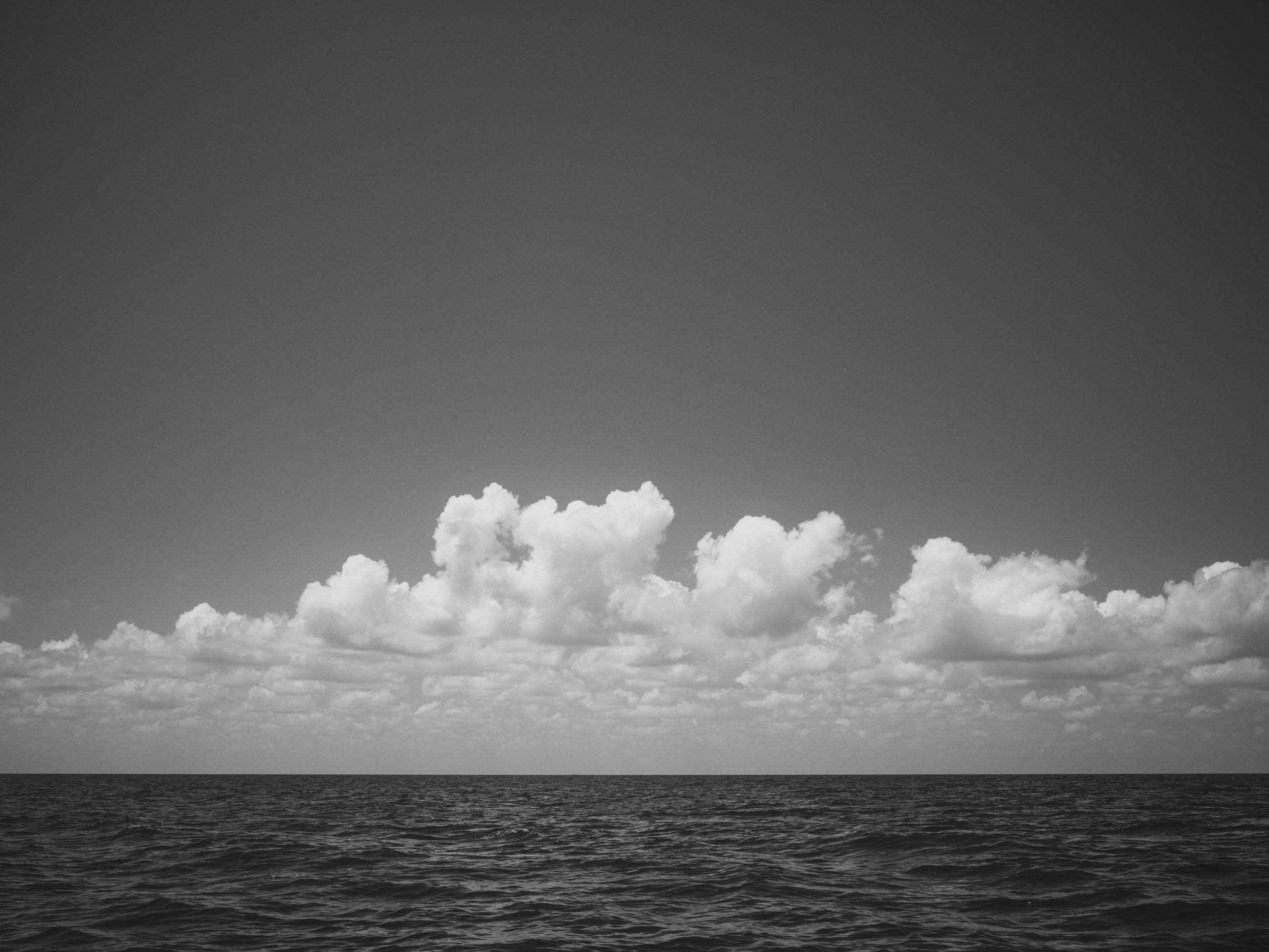 Black and white photograph of a seascape with water in the foreground, and a vast sky filled with clouds in the background.