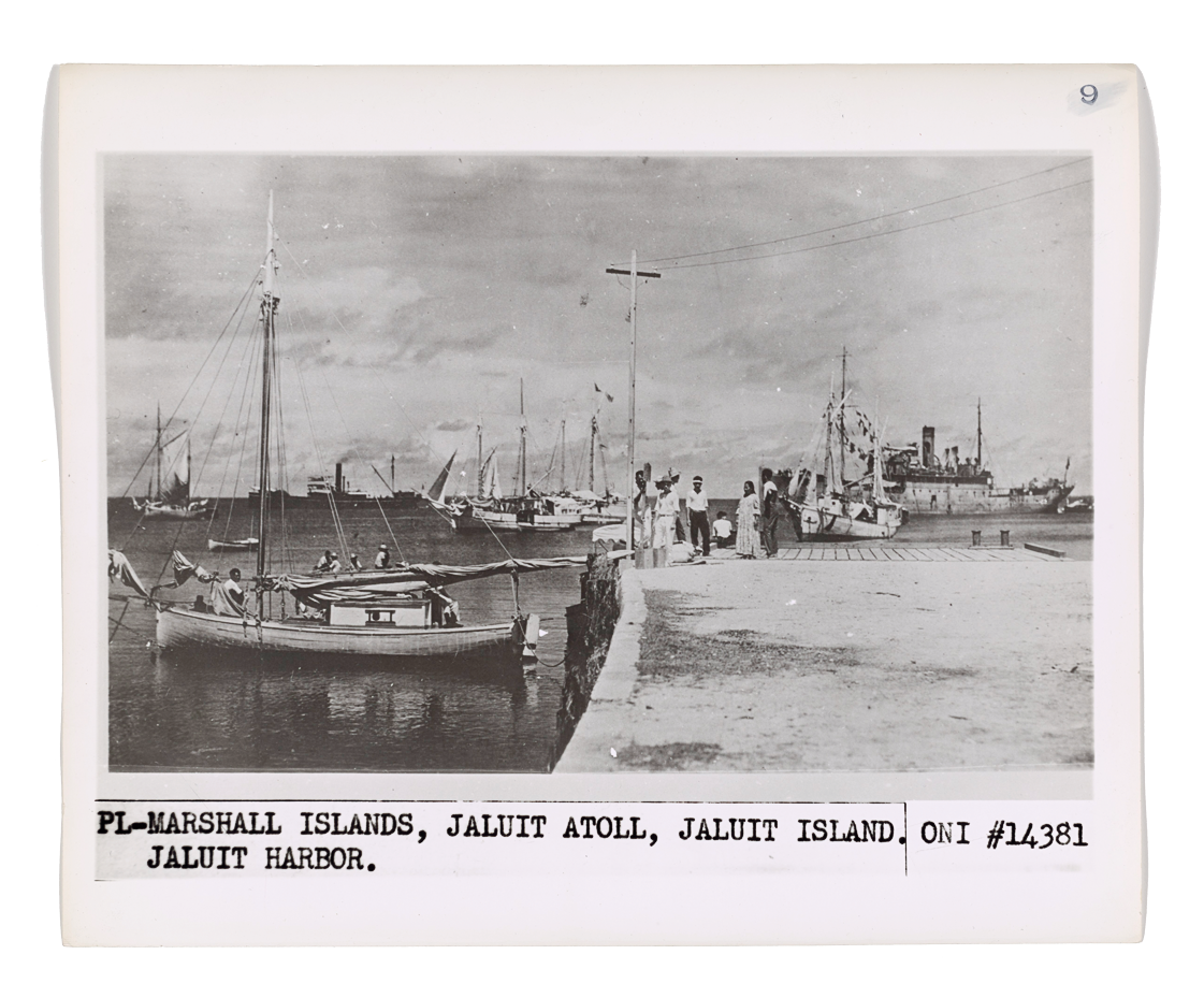 Black and white photo of Marshall Islands, Jaluit Atoll, Jaluit Island, Jaluit Harbor with boats docked at the pier and people standing on the dock, with ships in the background.