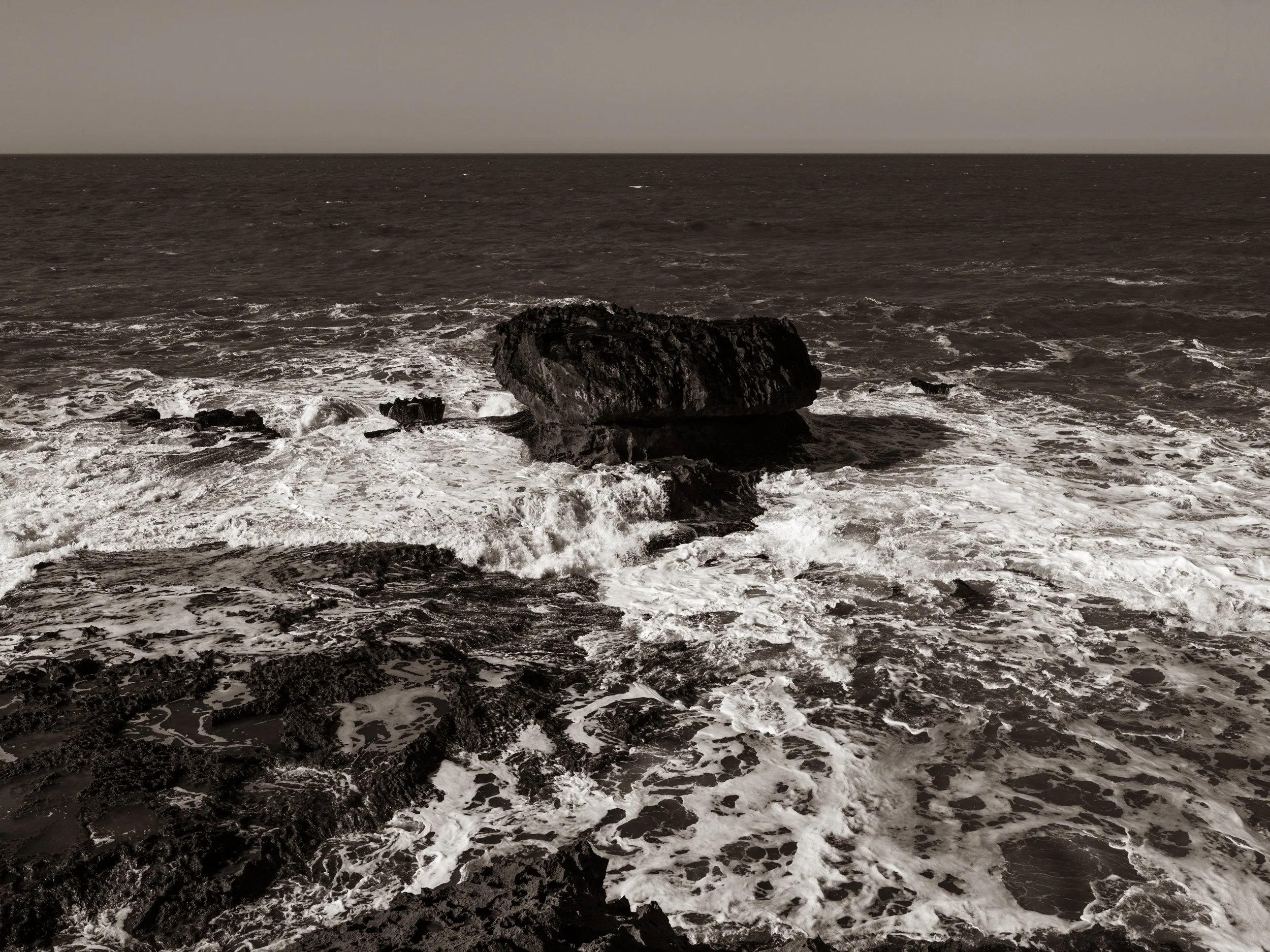 View of rough ocean waves crashing against dark rocks on the shoreline, with a cloudy sky in the background.