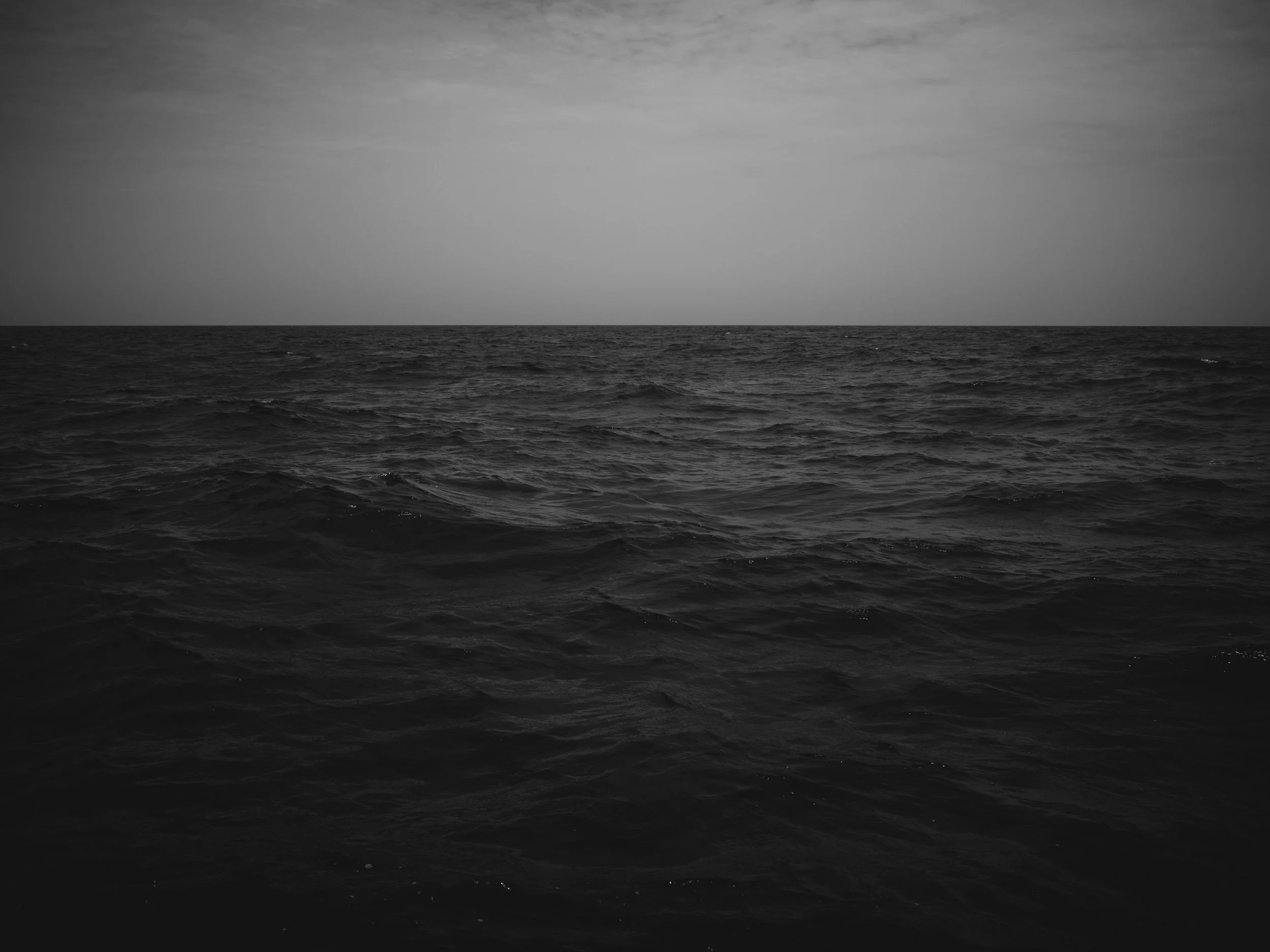 A black and white photo of the ocean with a horizon line where the water meets the sky, featuring textured waves in the foreground and wispy clouds in the sky.