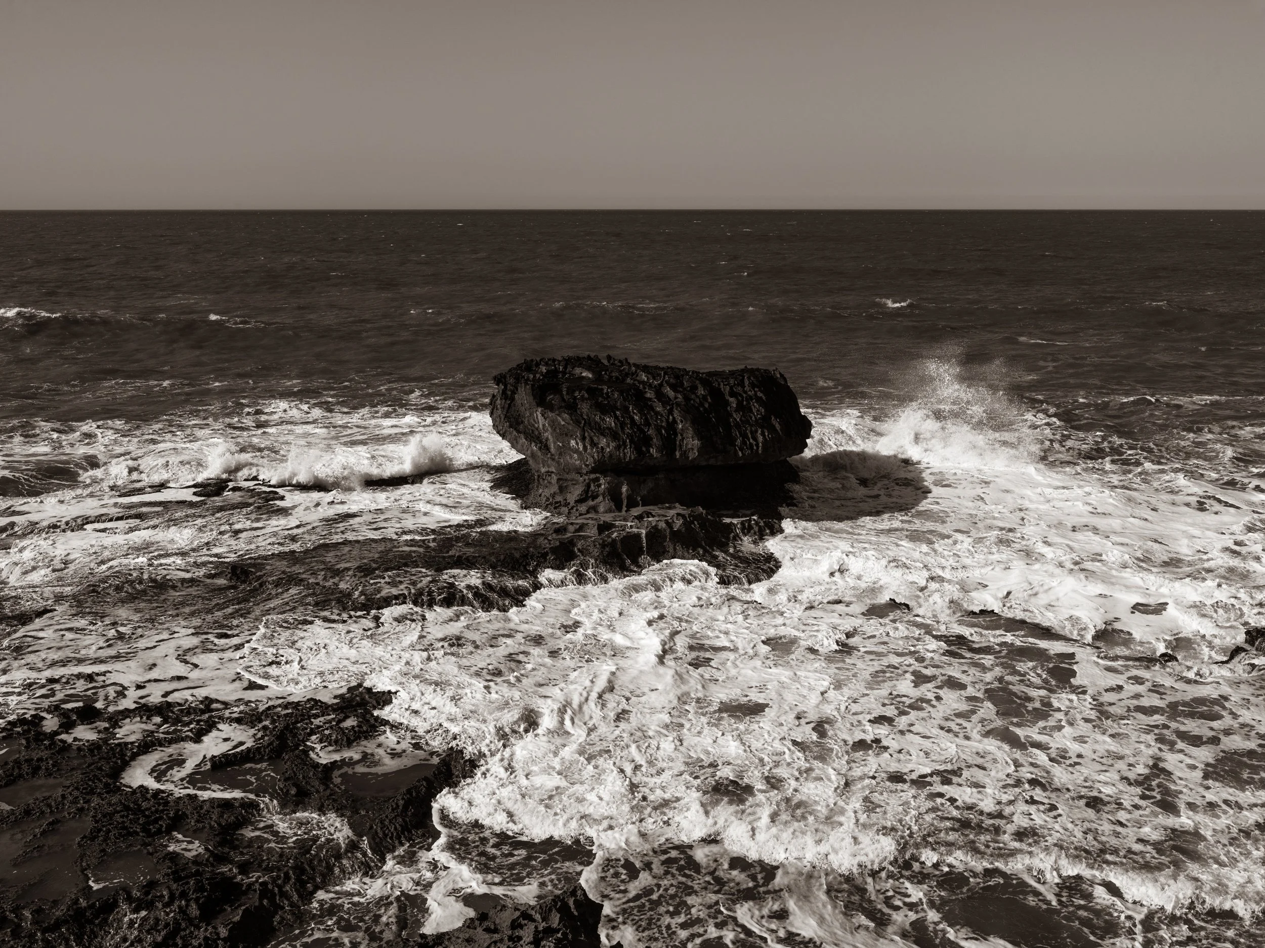 Black rock in ocean with waves crashing against it, cloudy sky in the background.