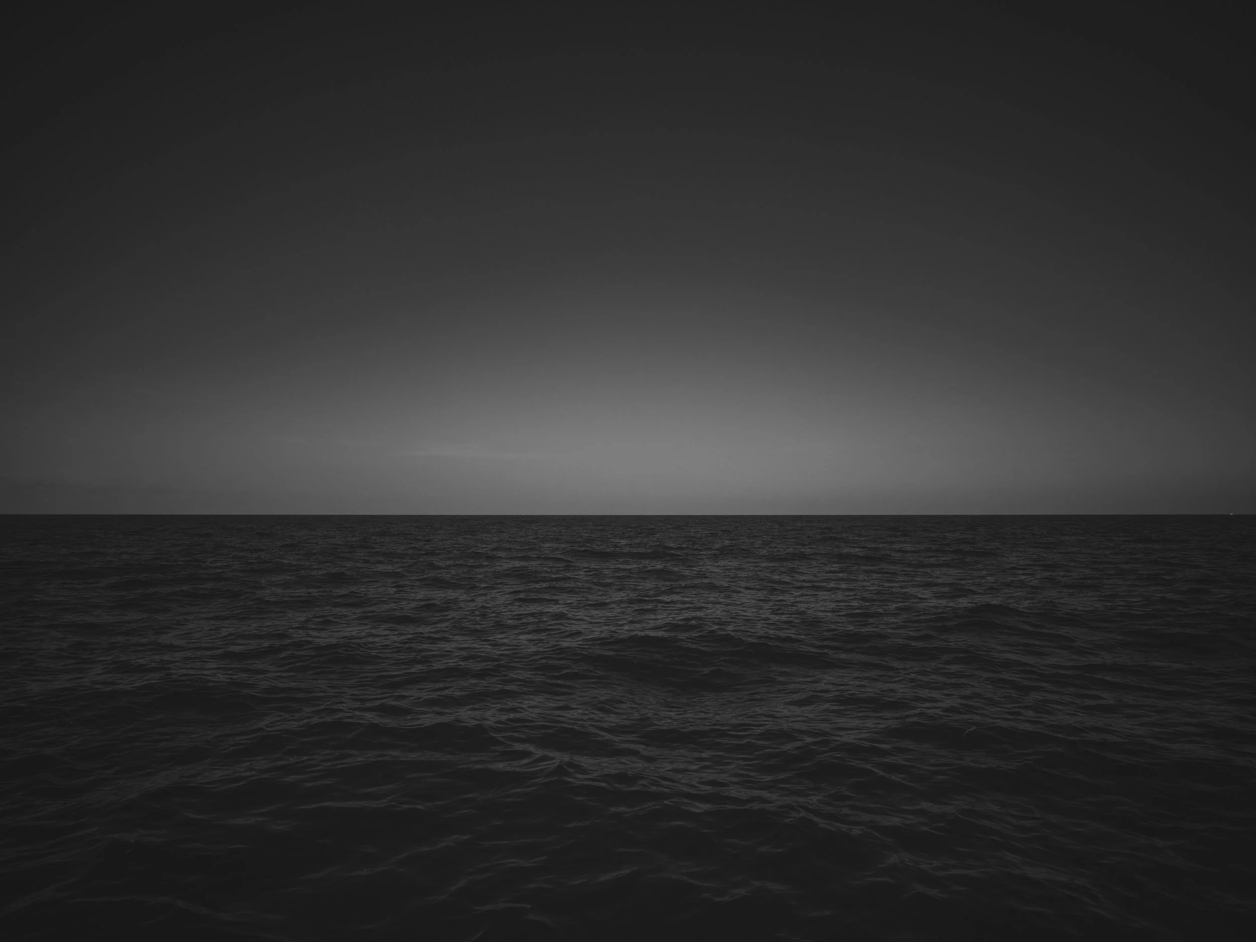 A black and white photograph of the ocean with a distant horizon and a dark sky.