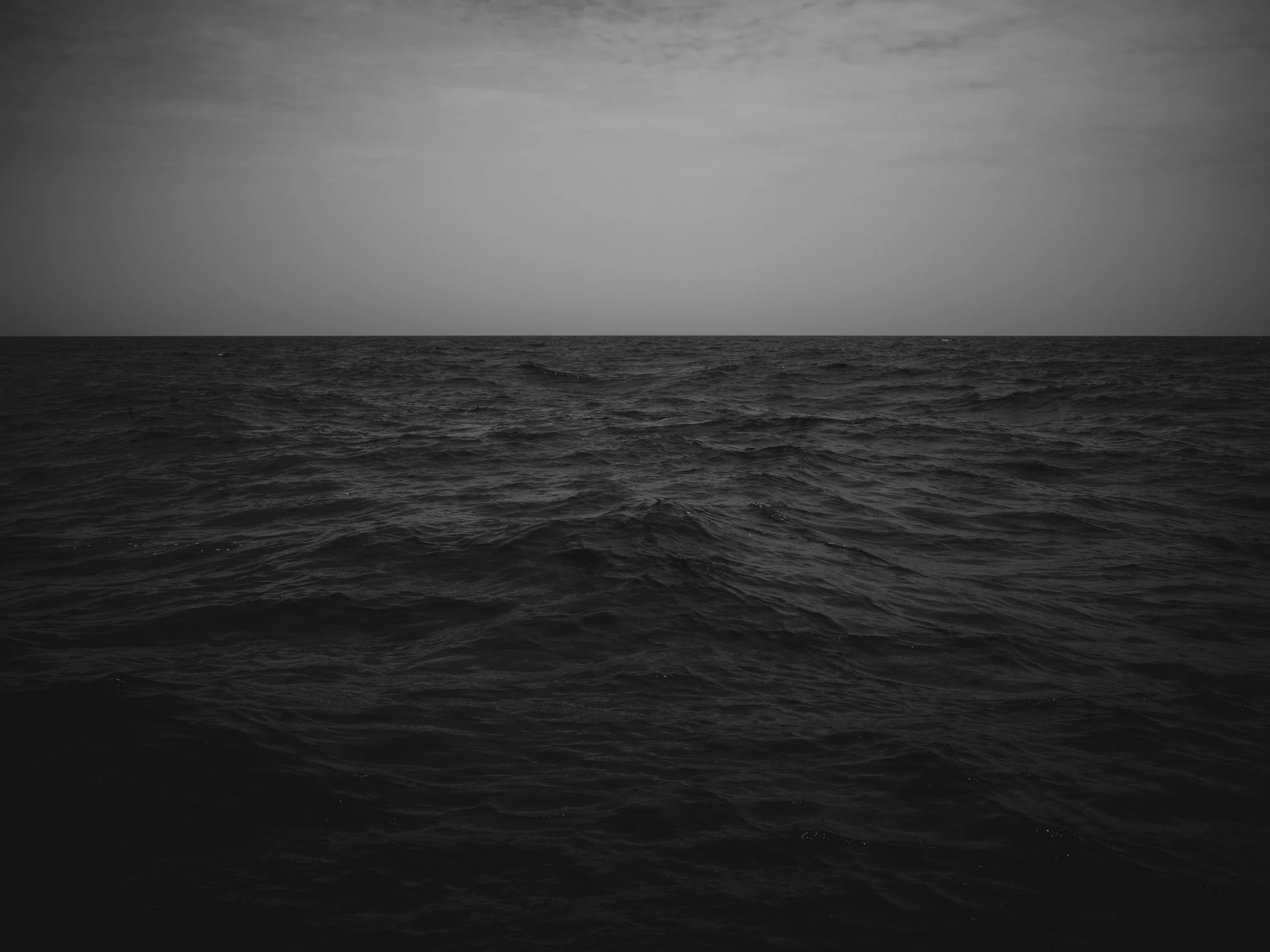 Black and white photo of an ocean with calm waves, under a cloudy sky.