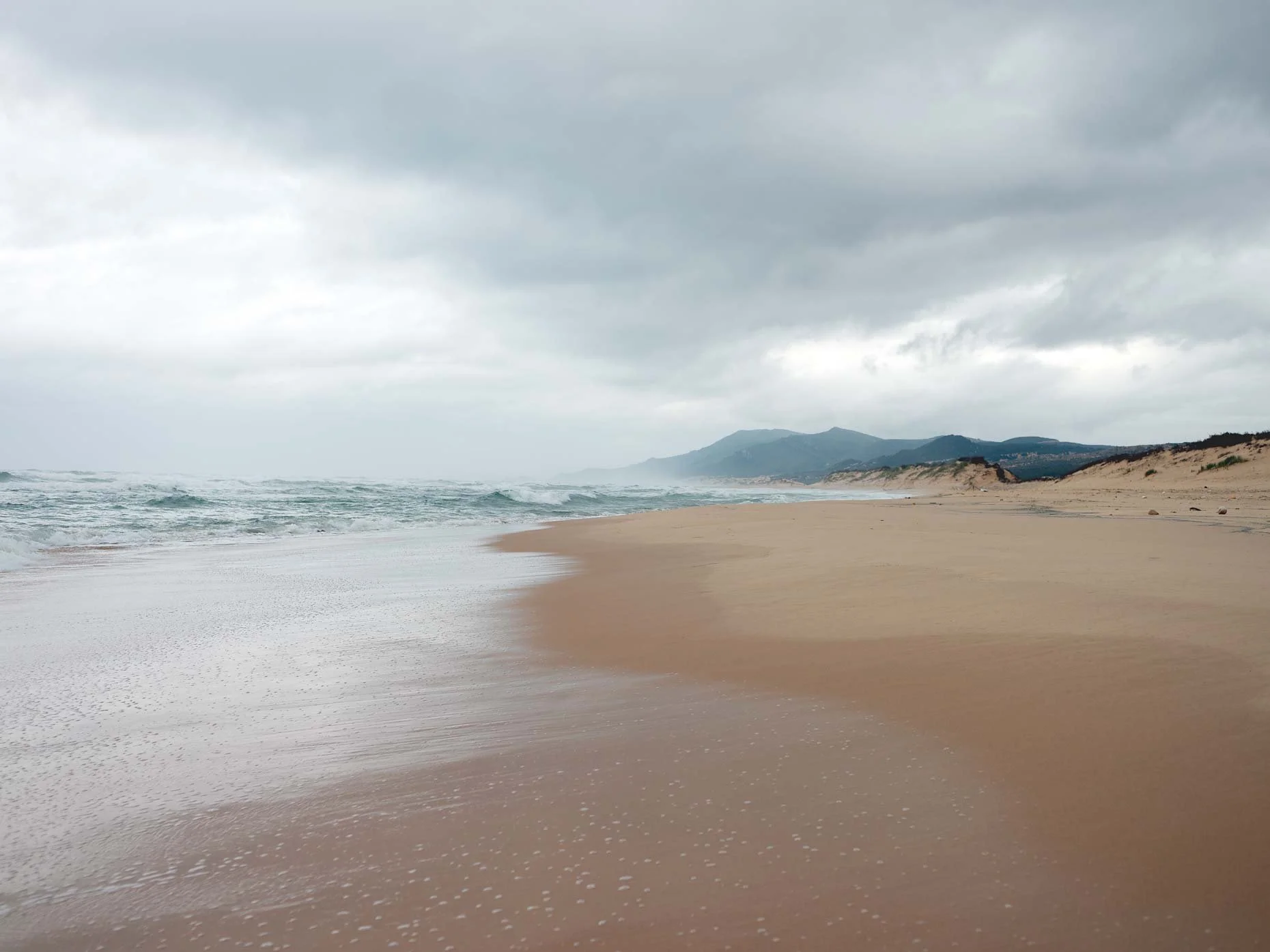 Empty sandy beach with gentle waves, distant mountains, and overcast sky.