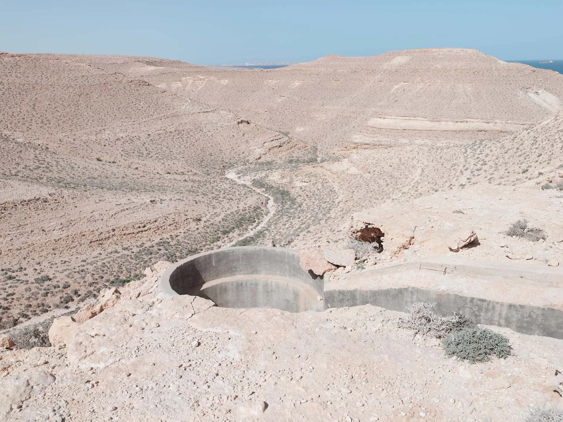 A barren, rocky desert landscape with sparse vegetation and rolling hills in the distance. In the foreground, there is a large, circular concrete structure embedded in the ground, possibly an industrial or water management feature.