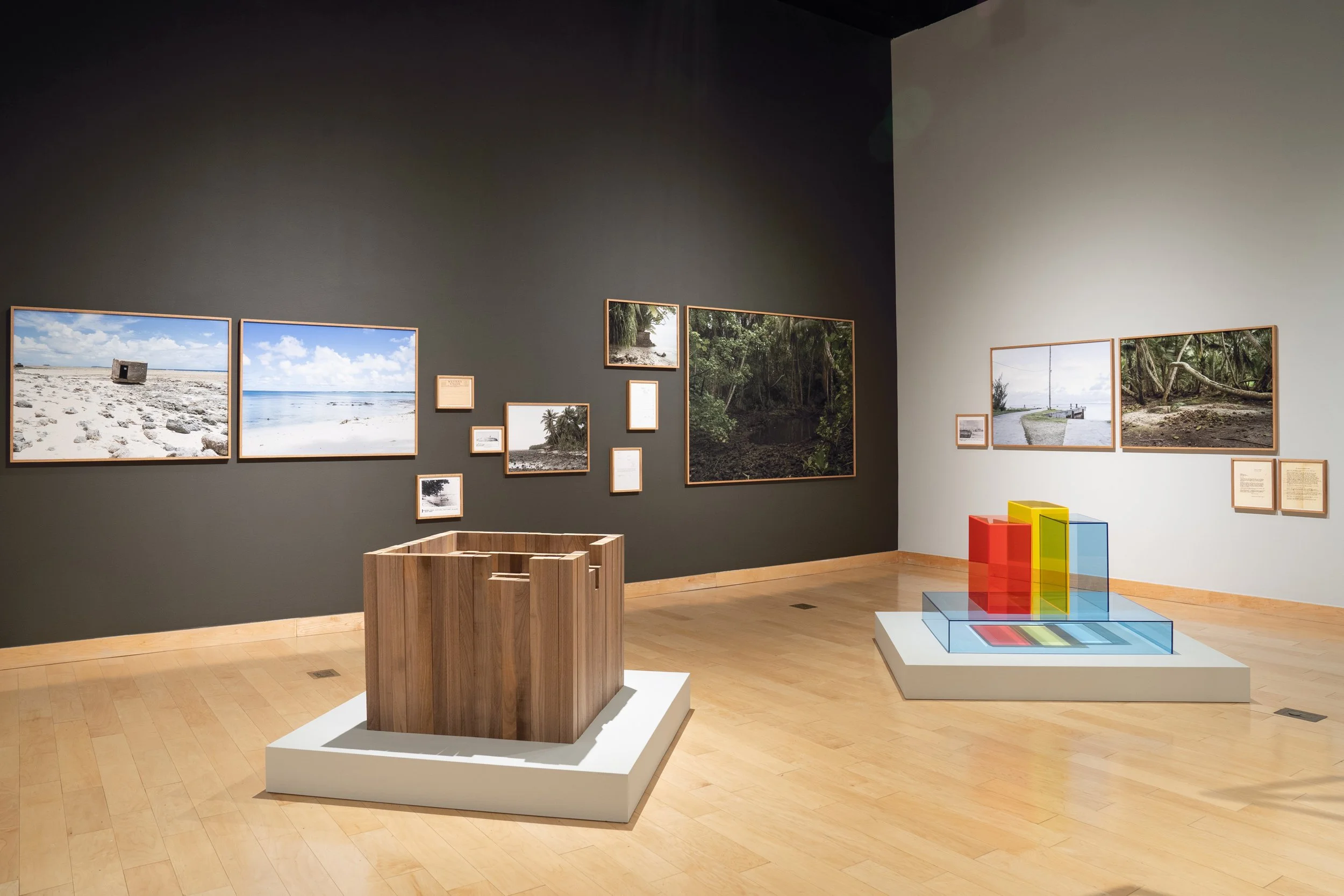 Art gallery with landscape photographs on dark and light gray walls, wooden and white display furniture, and colorful geometric sculpture.