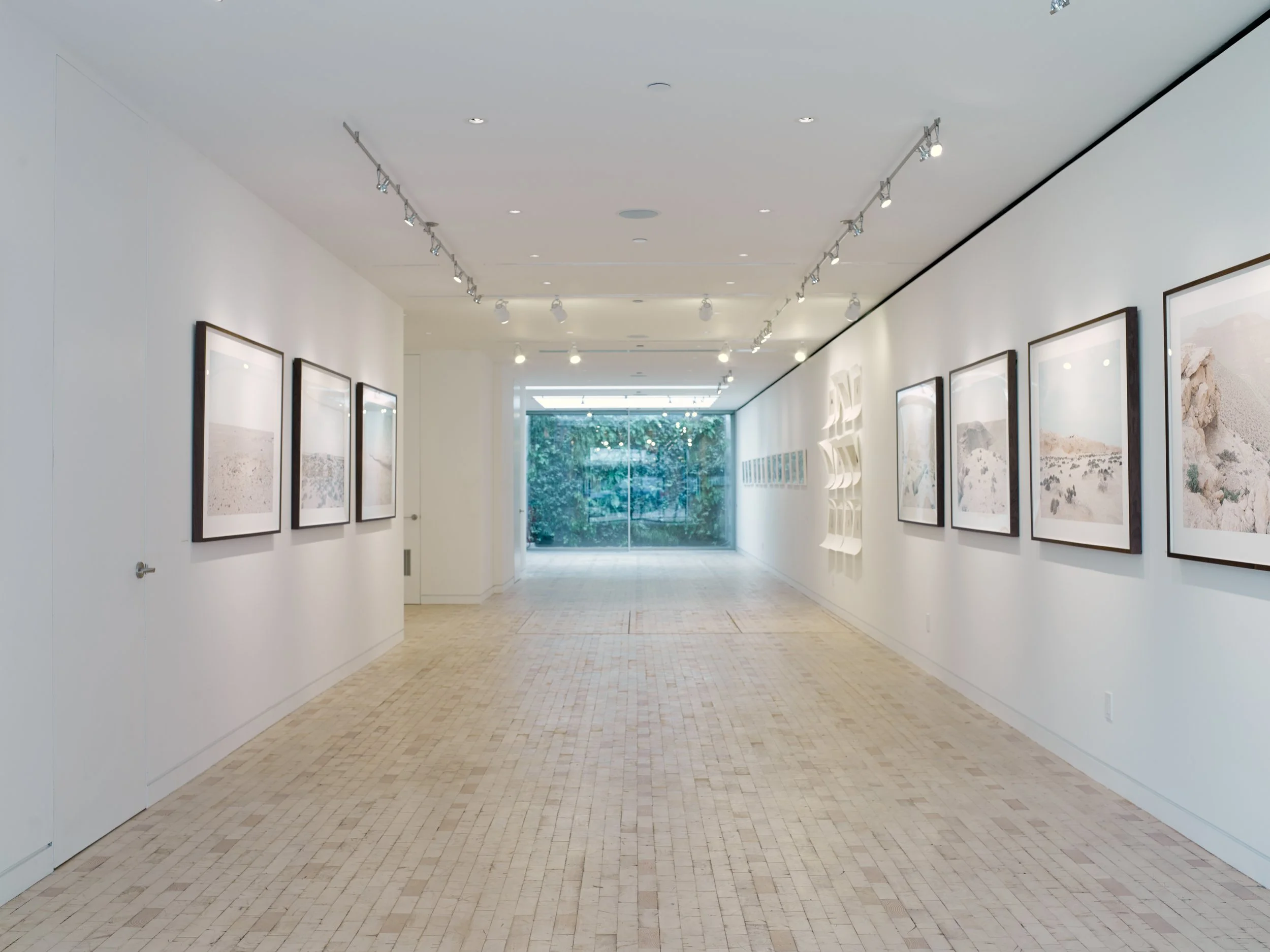 An art gallery with framed pictures on white walls, track lighting on the ceiling, a large window at the end with greenery outside, and beige tiled flooring.