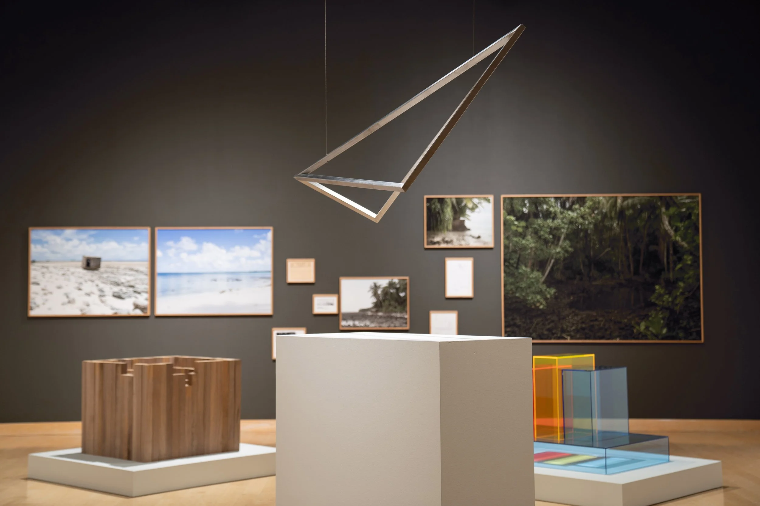 Art gallery interior with framed landscape photographs on dark wall, wooden and white display pedestals, and modern geometric sculpture hanging from ceiling.
