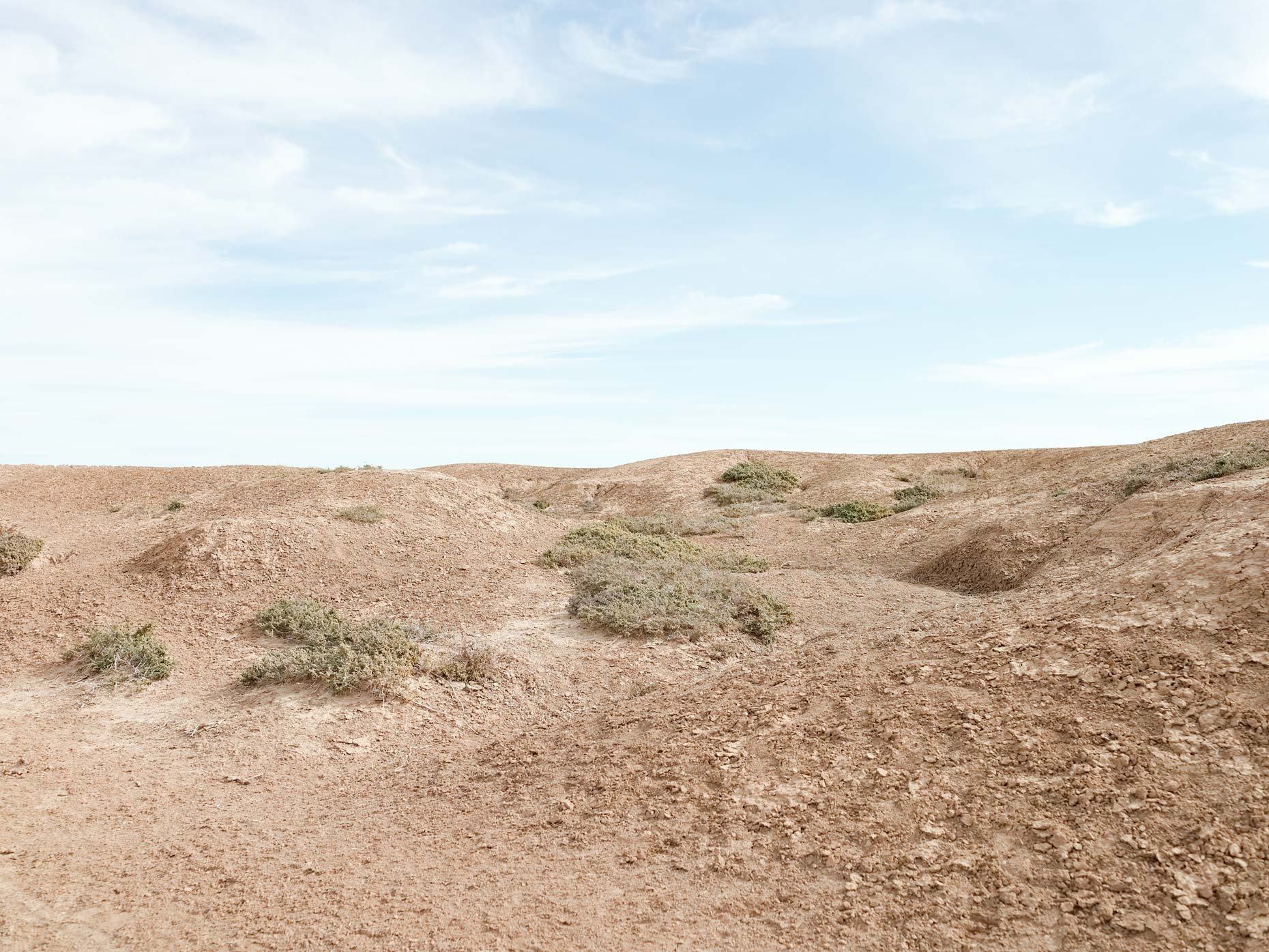 Arid desert landscape with sandy terrain and sparse green shrubs under a partly cloudy sky.