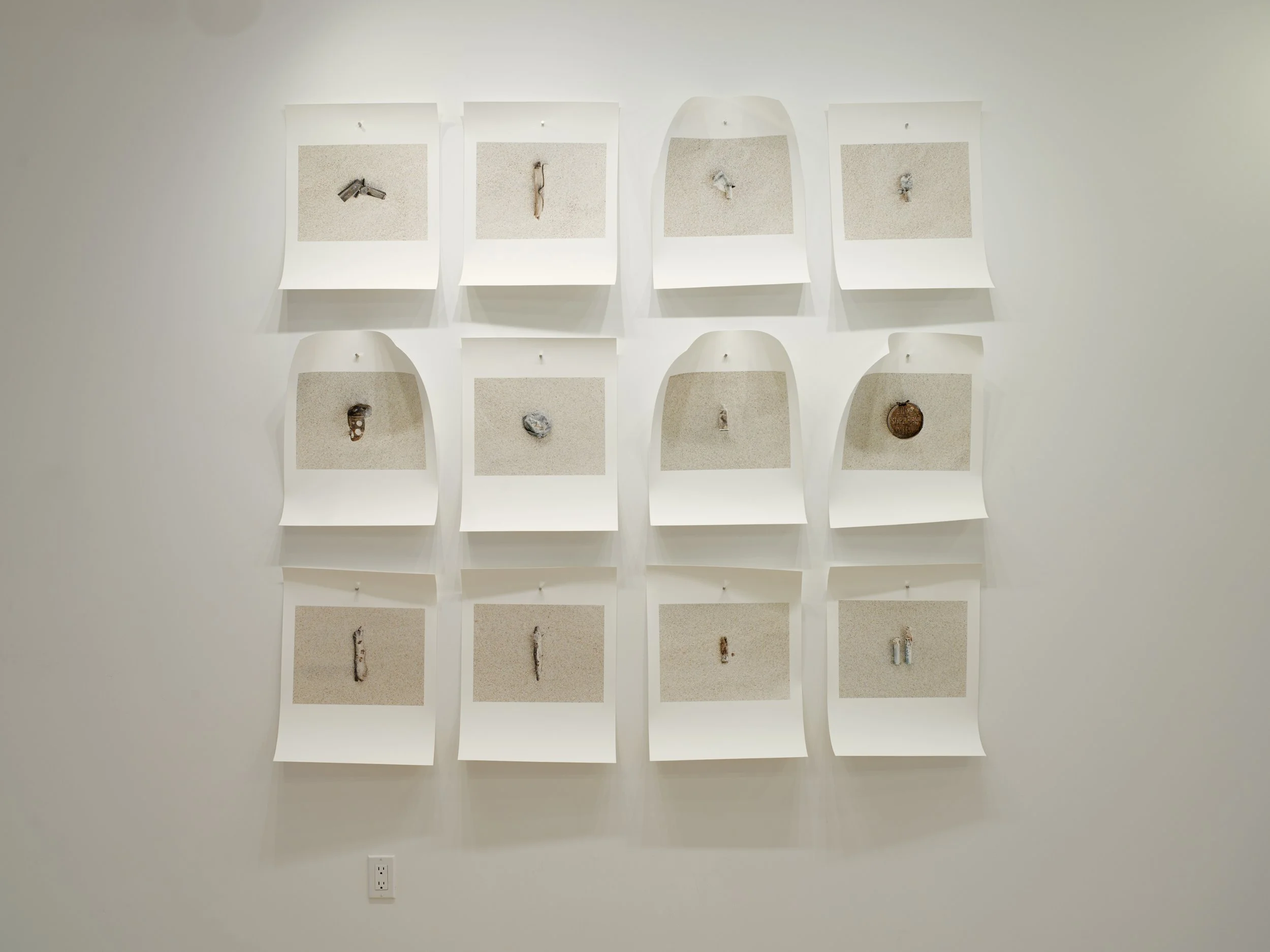 A wall with nine small photographs arranged in a 3x3 grid, each depicting a different object on a sandy surface, some partially obscured or distorted.