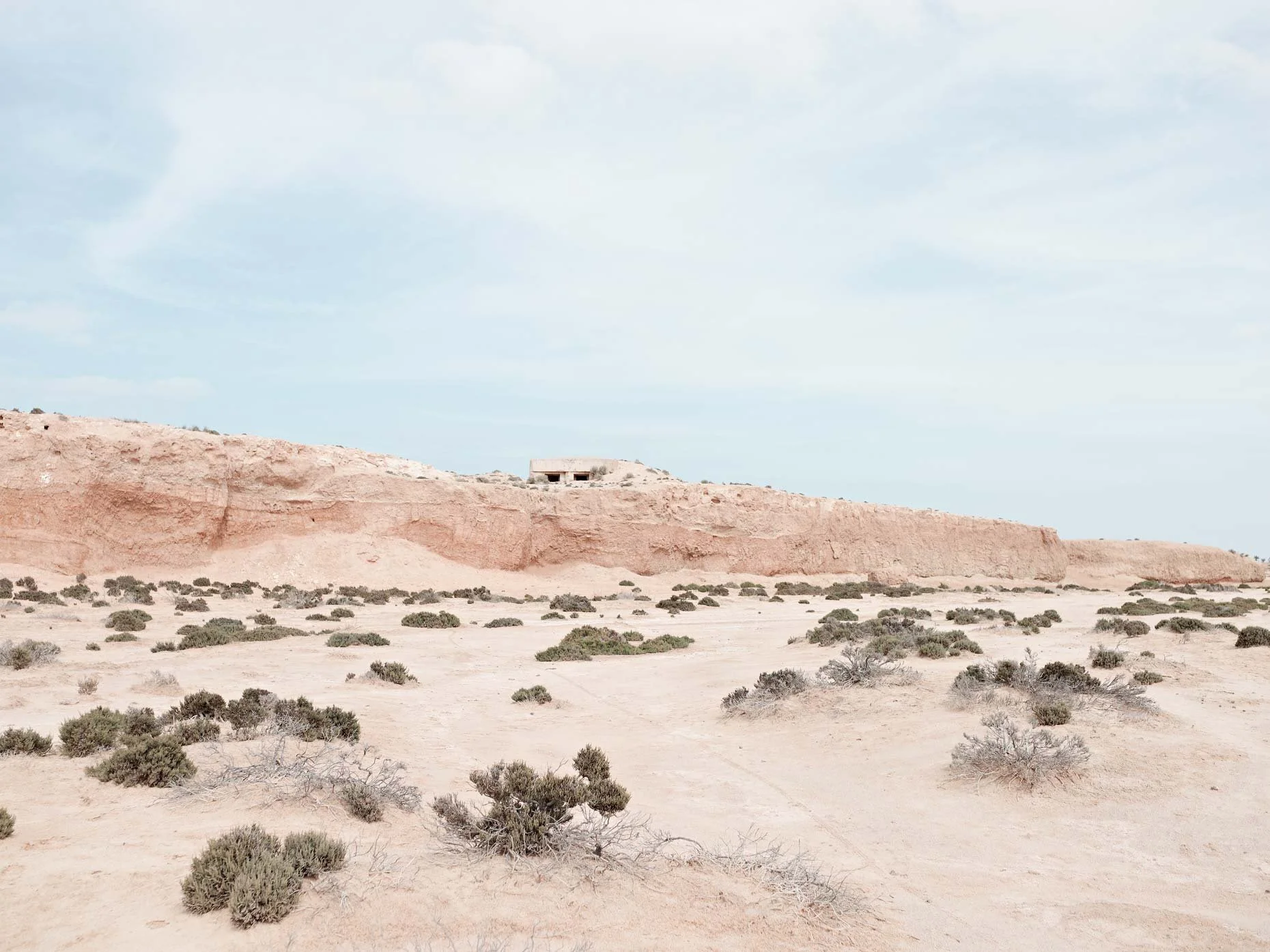 Desert landscape with scattered bushes, red cliffs in the background, and a small building atop the cliffs under a cloudy sky.