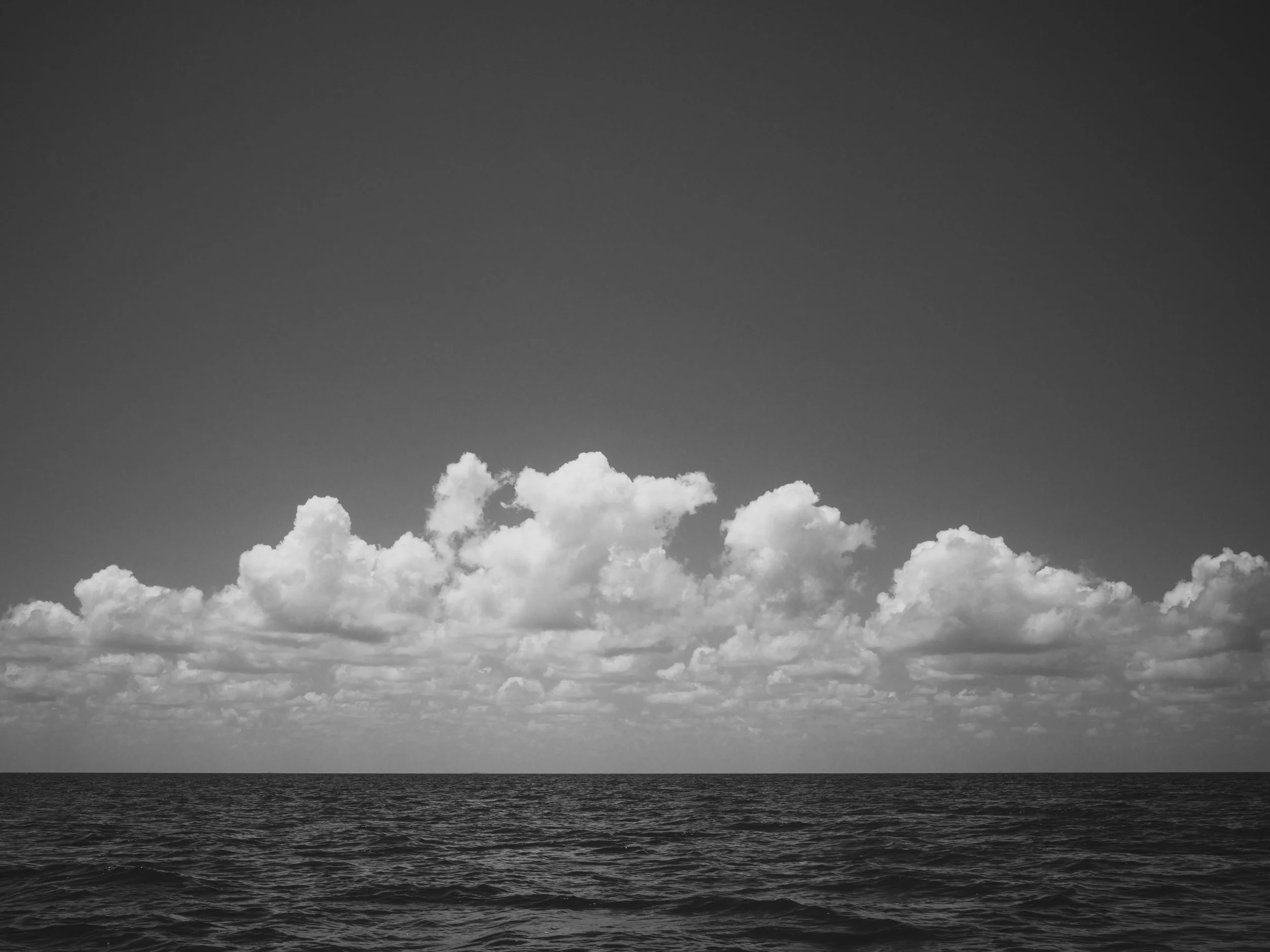 Black and white photo of the ocean with a clear horizon line, under a sky filled with scattered clouds.