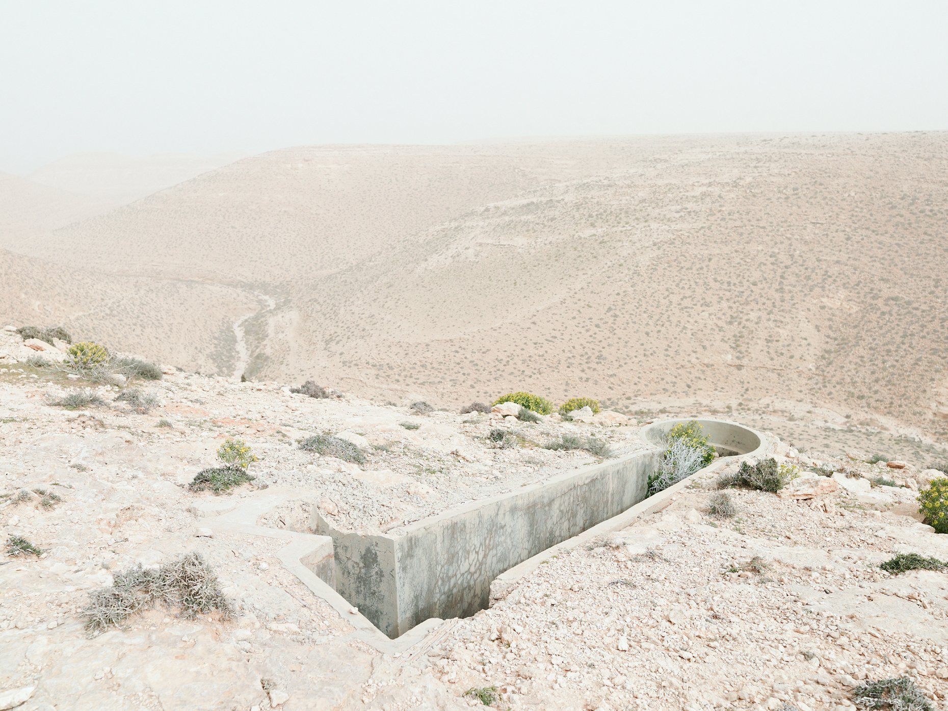 Desert landscape with sparse vegetation and a man-made concrete water trough or drain in the foreground, with rolling sandy hills in the background.