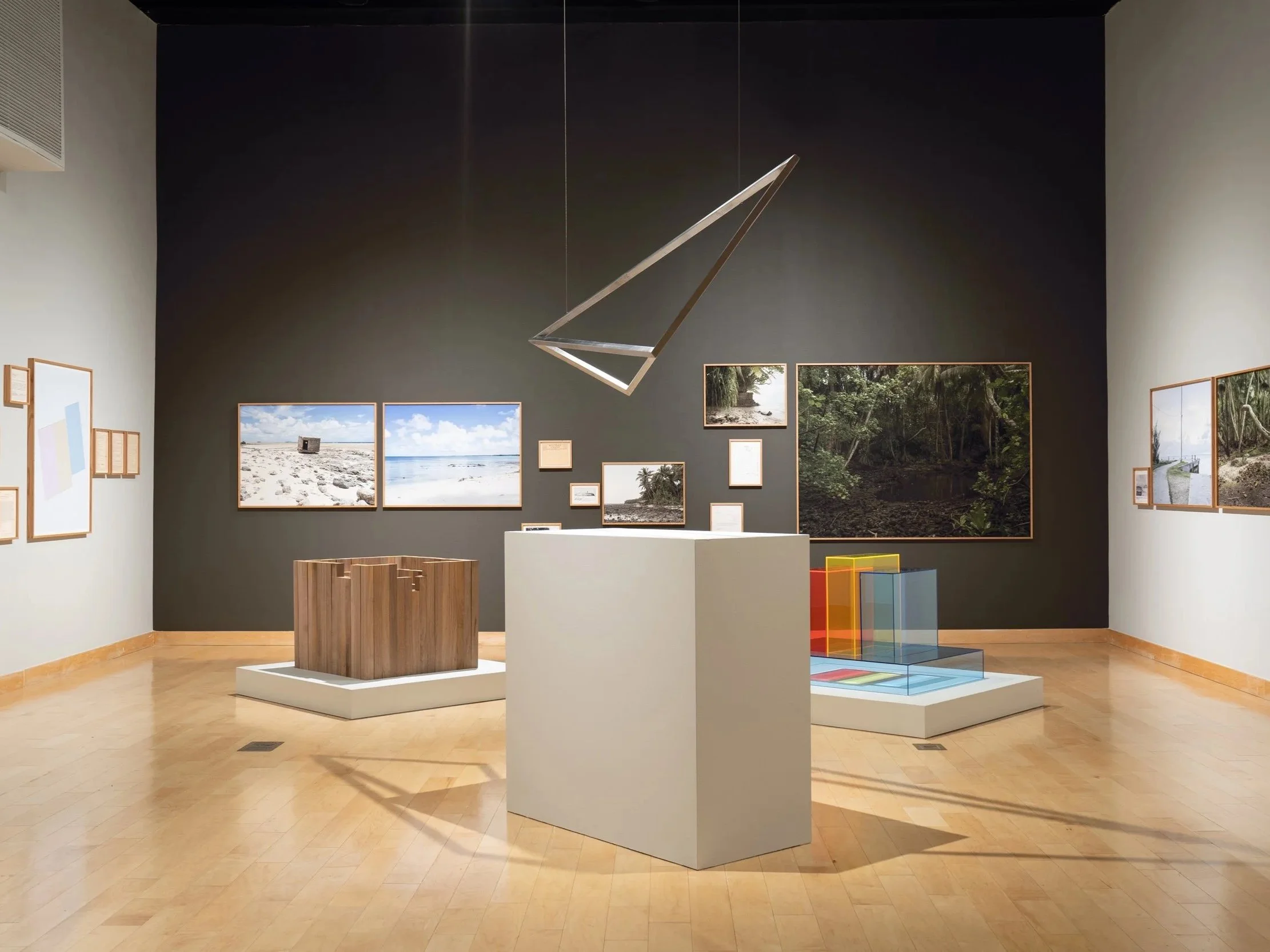 Art gallery exhibit with various framed photographs and artworks on gray walls, including beach scenes, forest images, and architectural photos. White pedestals display sculptures, with abstract colorful glass pieces on the right. A modern rectangular light fixture hangs from the ceiling.