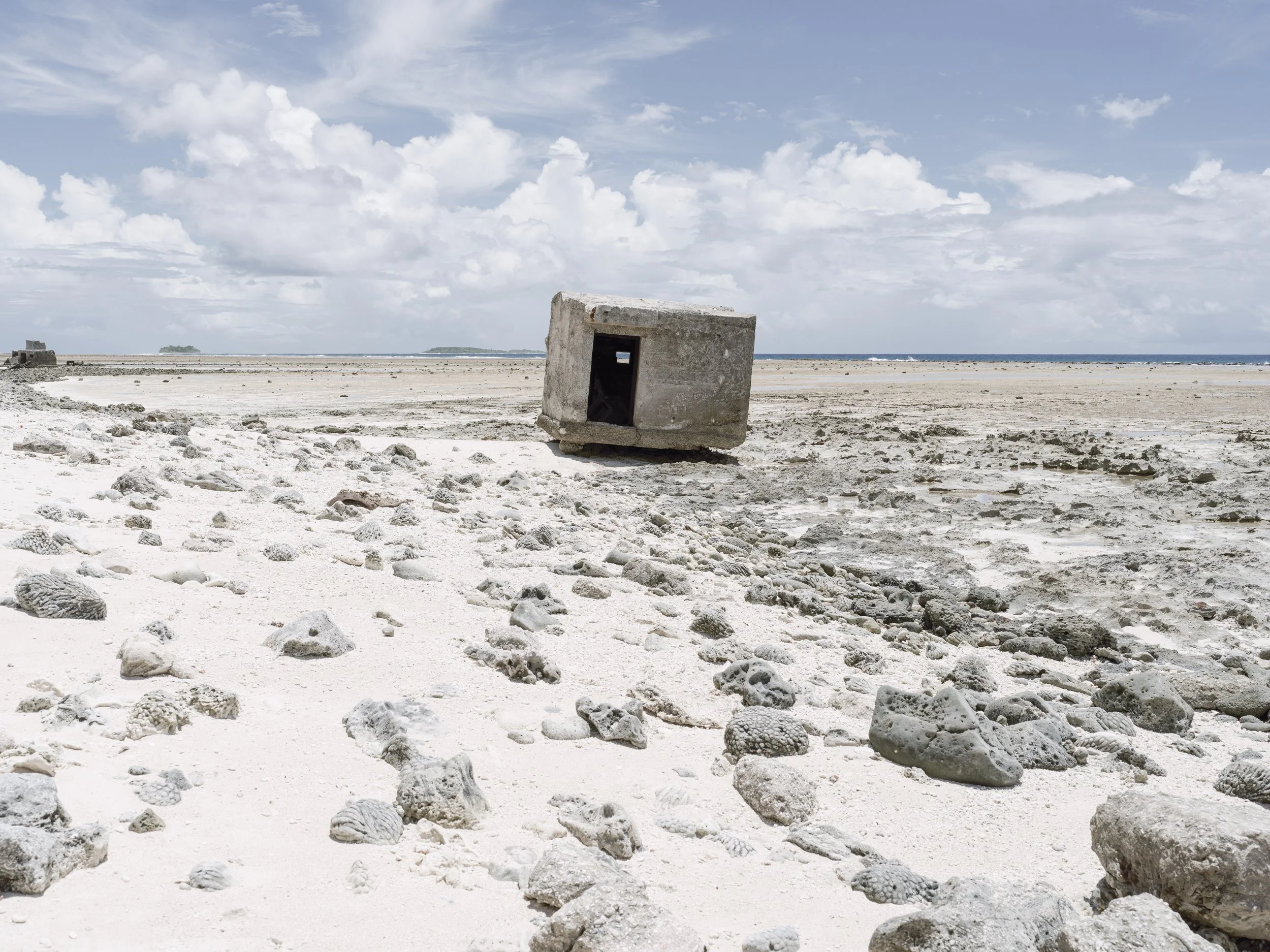 A sandy beach with scattered rocks and a small concrete structure with a rectangular opening, under a partly cloudy sky with some distant islands visible across the water.