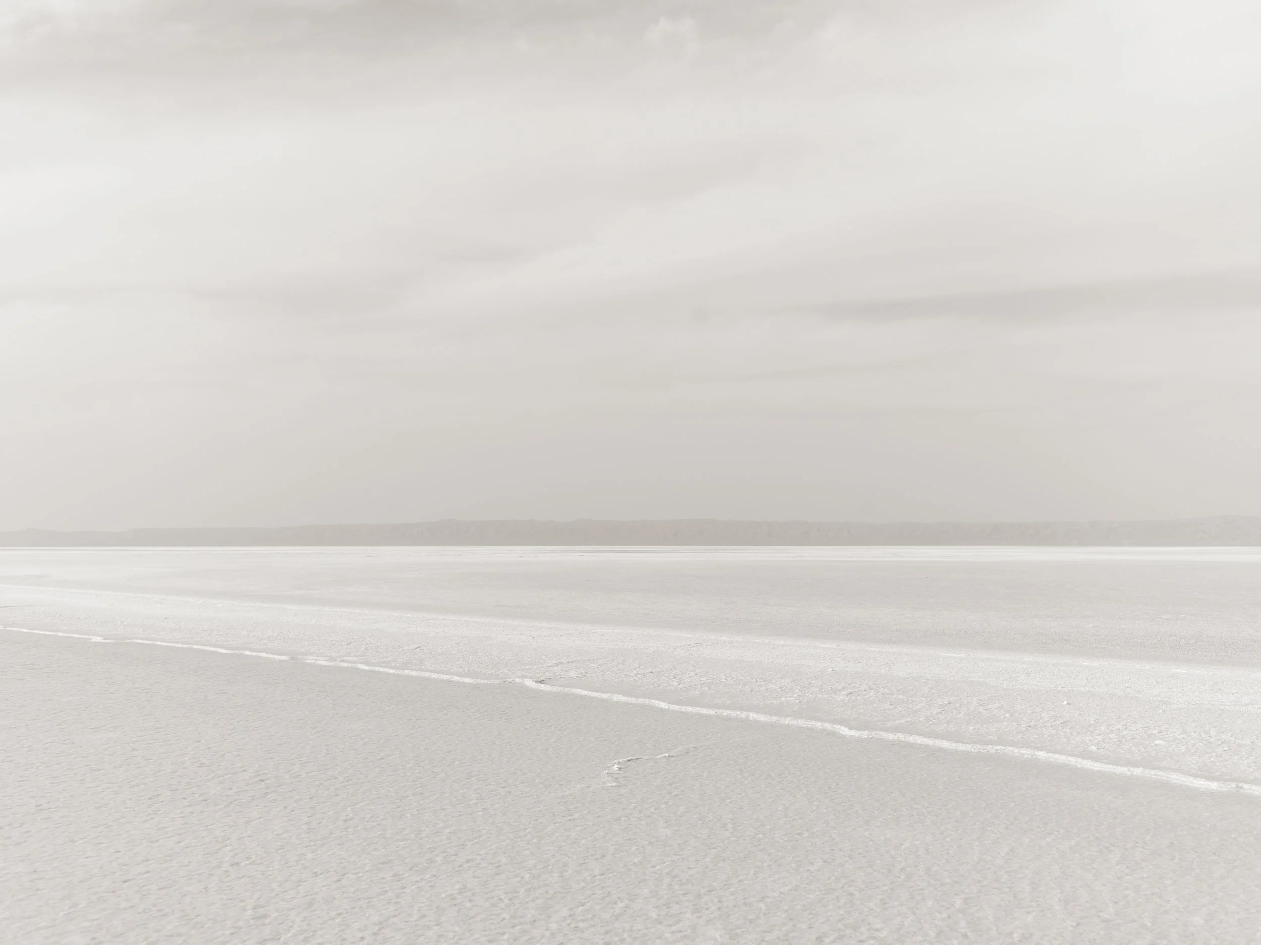 An expansive, flat salt flat with a white, textured surface. The sky is overcast with gray clouds, and there is a faint horizon line in the distance.