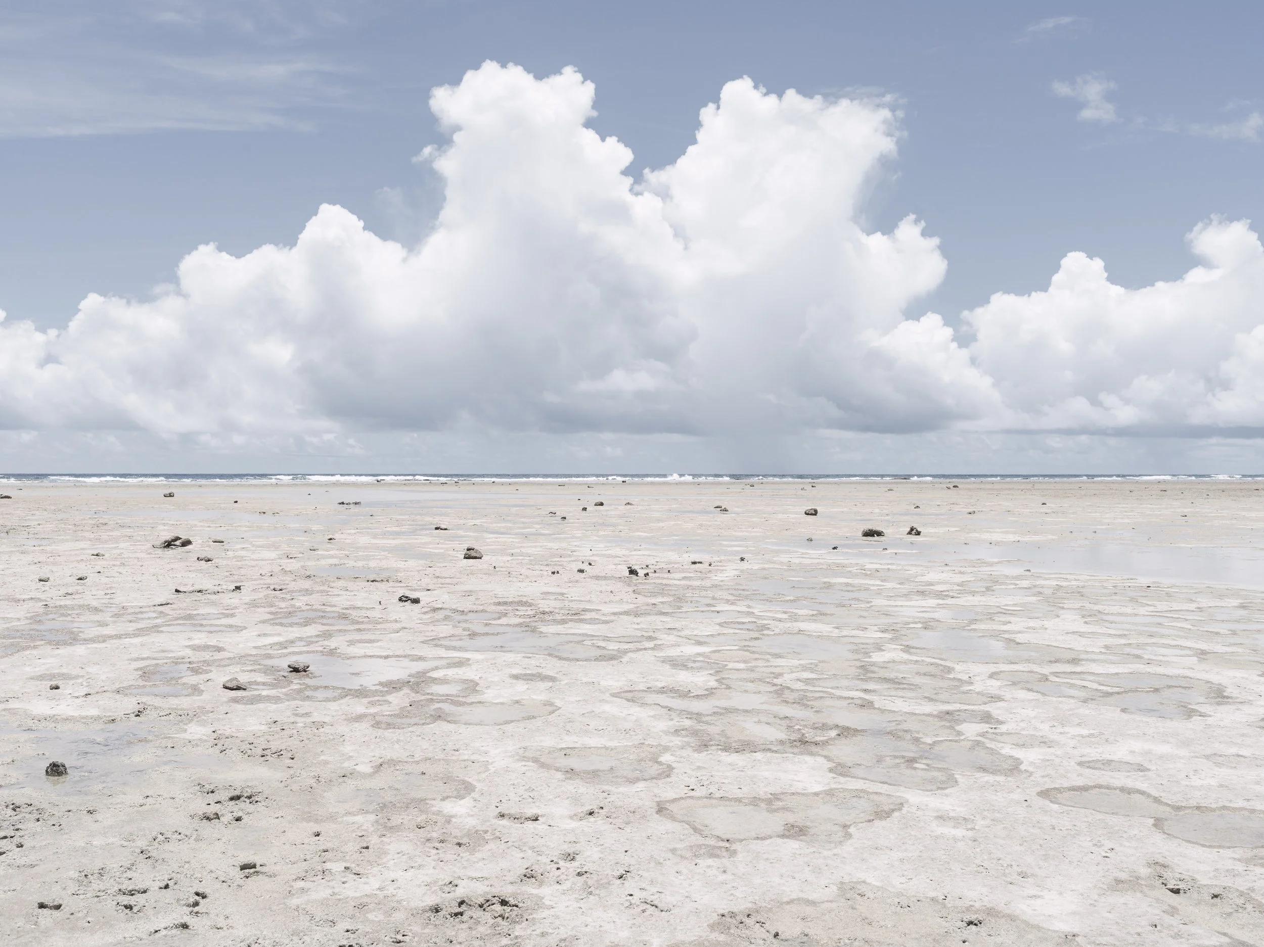 A sandy beach with stones scattered across the surface, under a blue sky filled with large, fluffy white clouds.