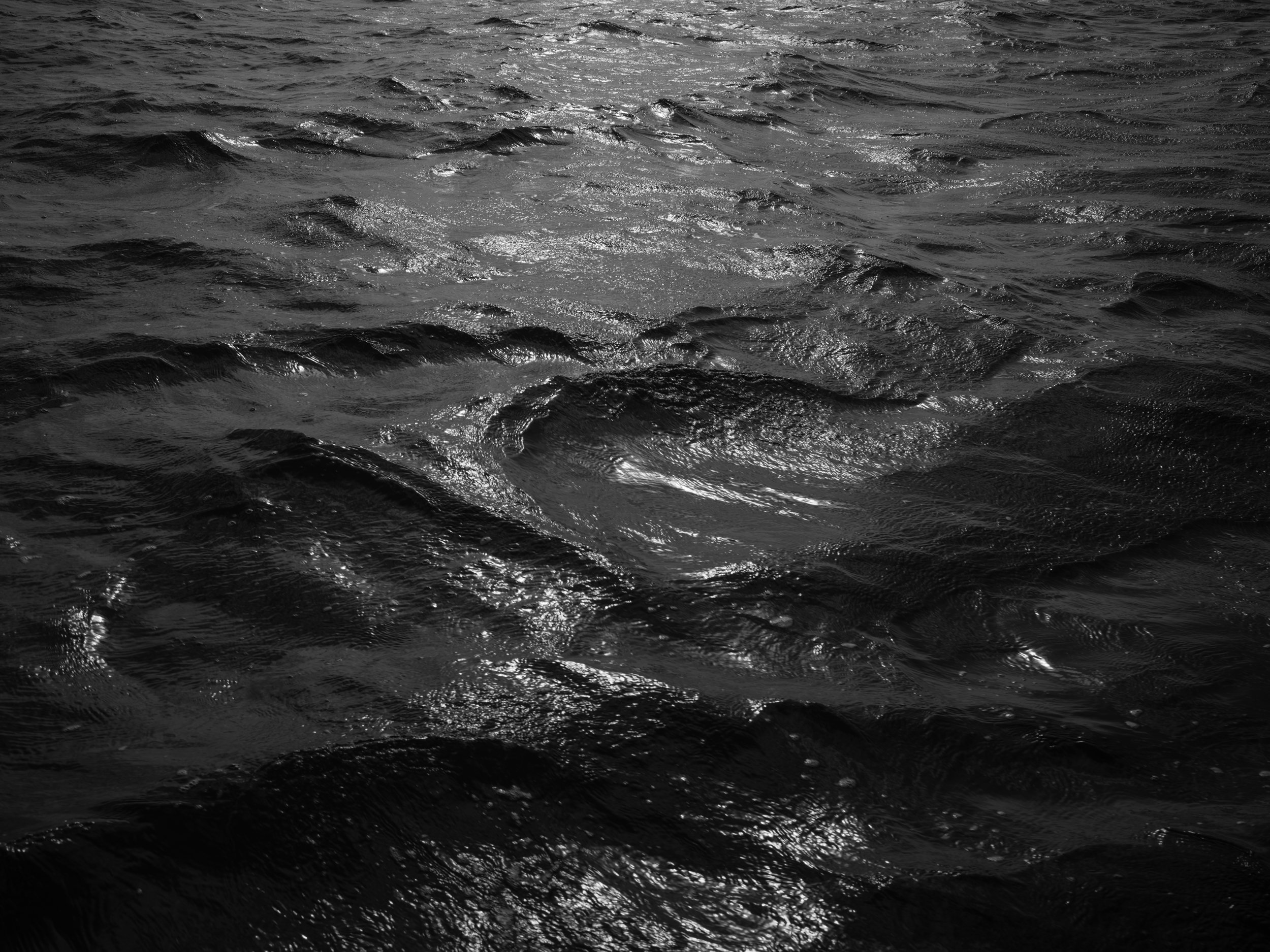 Black and white photo of ocean water with ripples and sunlight reflecting off the surface.