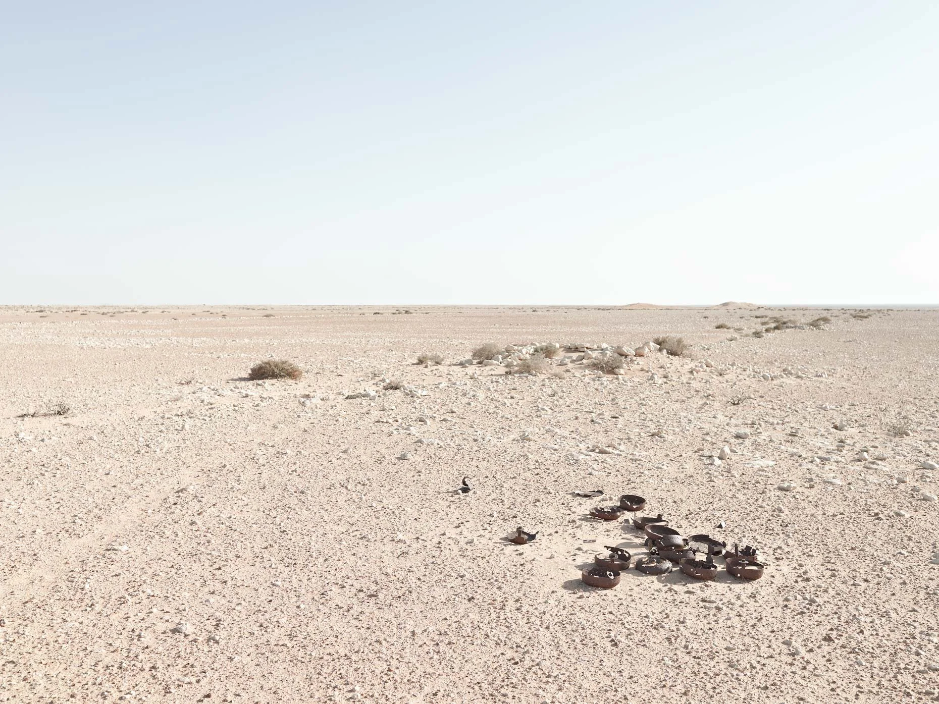 Desert landscape with sparse bushes and a clear sky, featuring a small pile of rusted metal objects in the foreground.