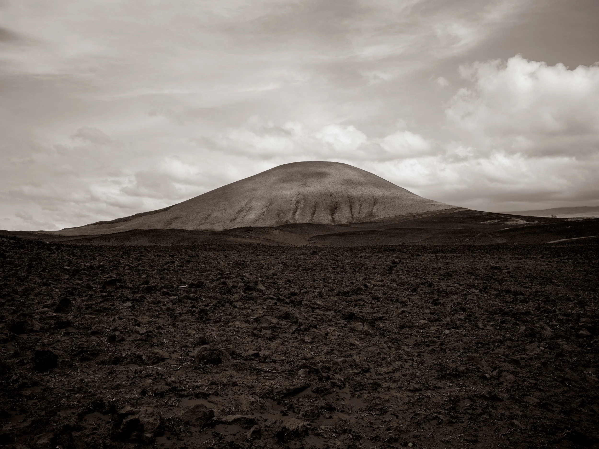 A lone, rounded hill or mountain with a smooth surface and faint lines running down its side, set against a cloudy sky, in black and white.