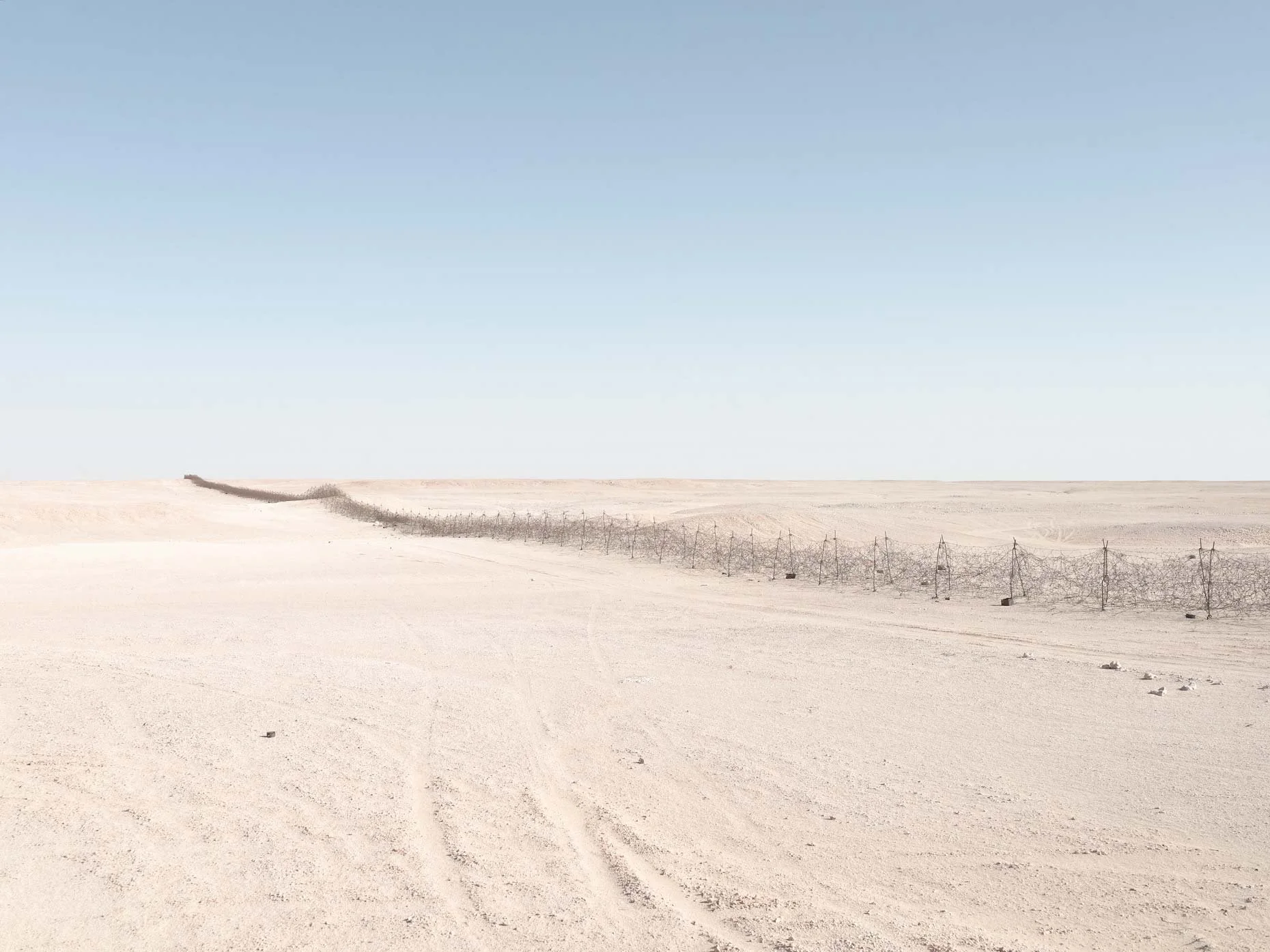 A vast, flat desert landscape with dry, light-colored sand and minimal vegetation, a barbed wire fence running through the scene, and a clear blue sky overhead.