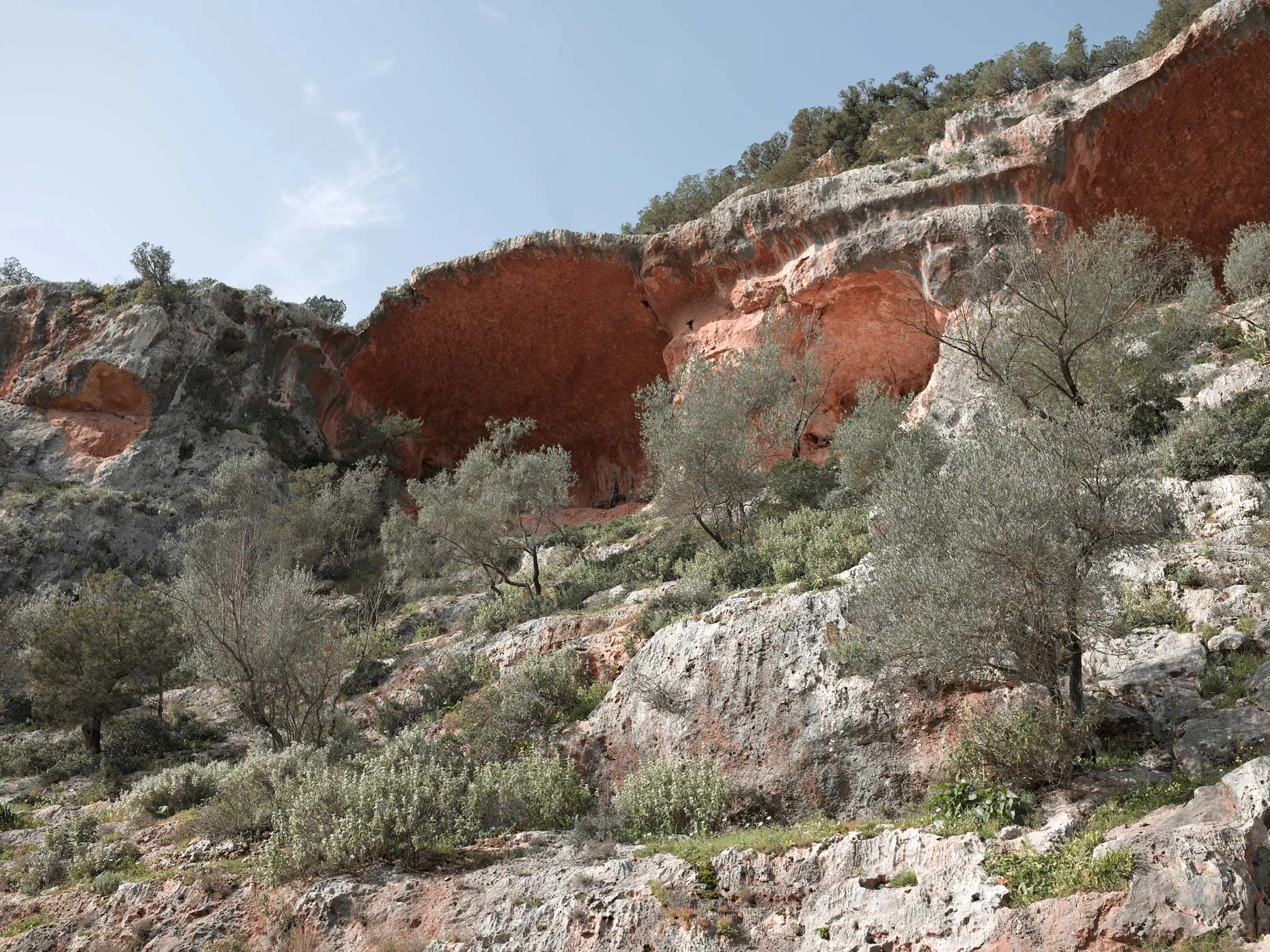 A rocky hillside with sparse vegetation and large red and gray rock formations under a clear blue sky.