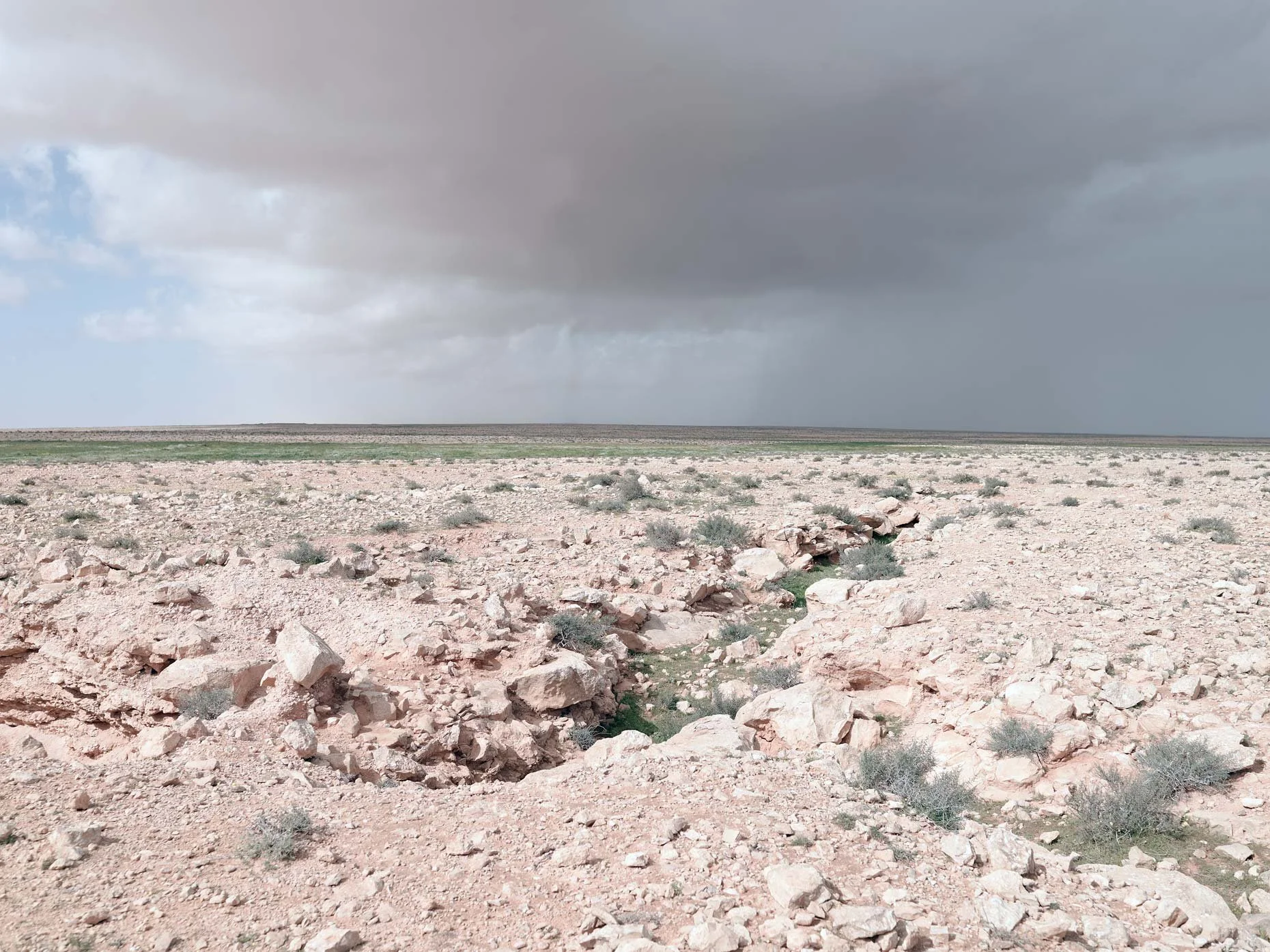 A barren desert landscape with scattered rocks and sparse vegetation, under a cloudy sky with dark, looming clouds.