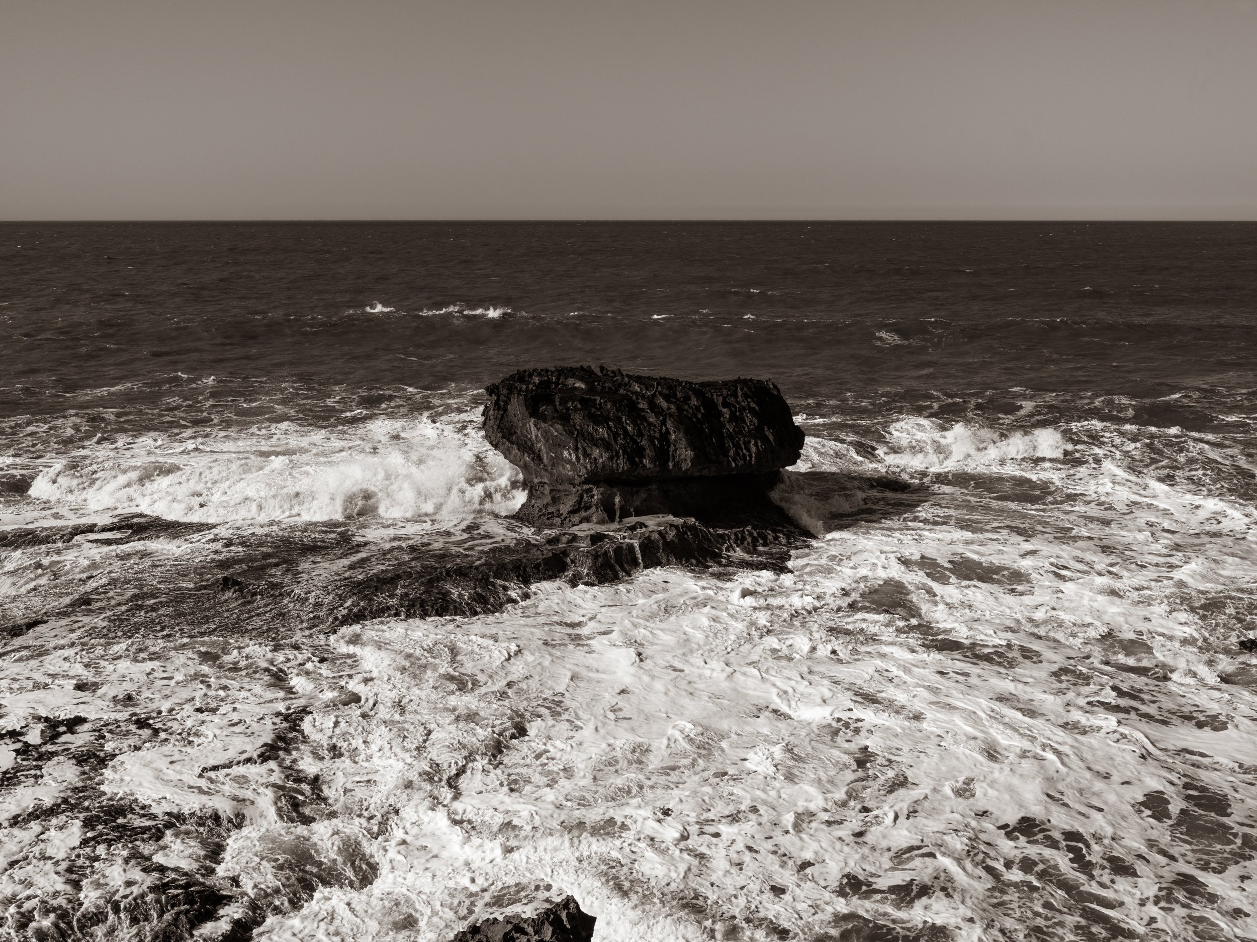 A large dark rock in the water near the shore, with waves crashing around it. The ocean extends to the horizon under a cloudy sky.