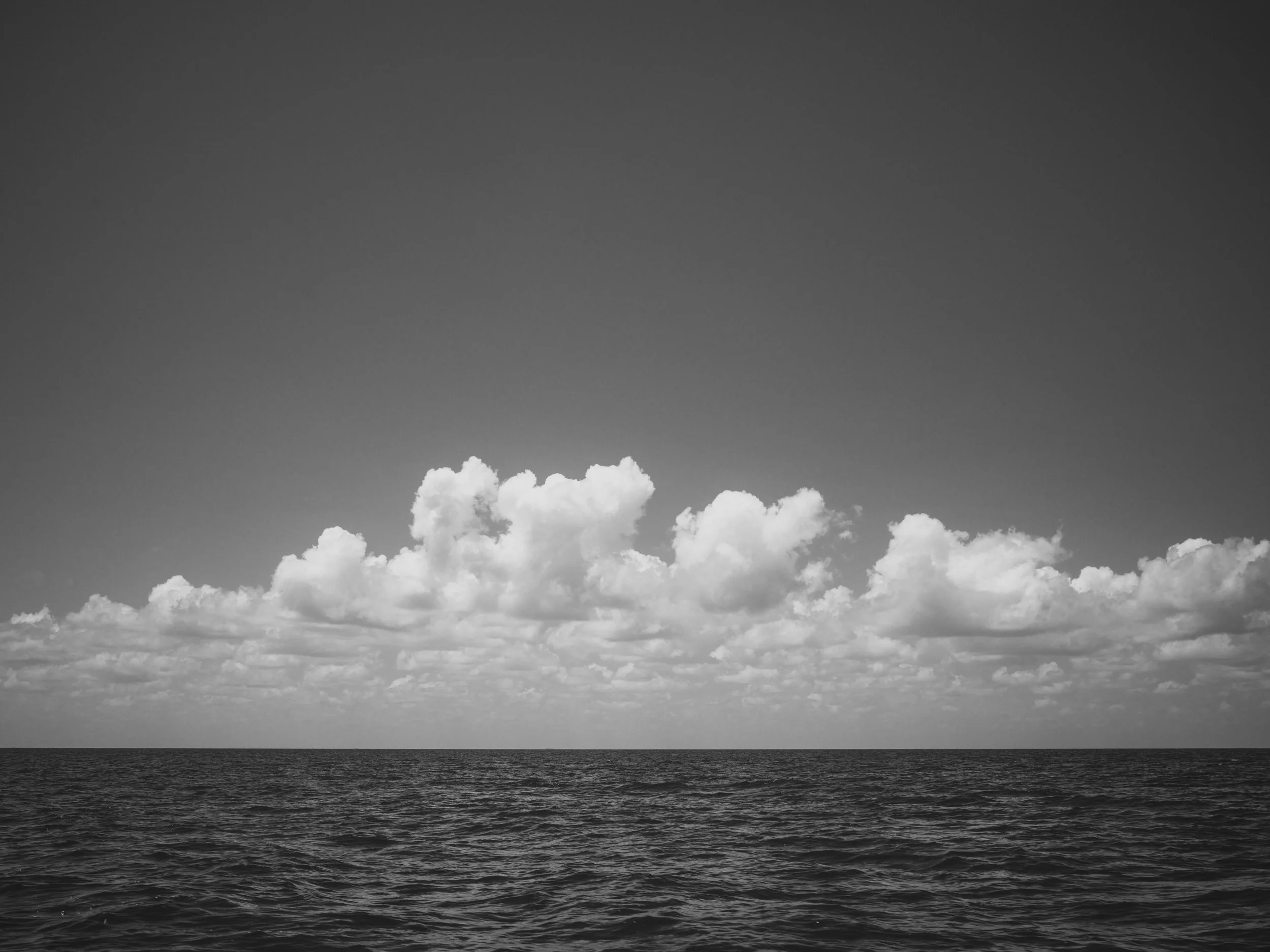 Black and white photograph of the ocean with a line of clouds in the sky.