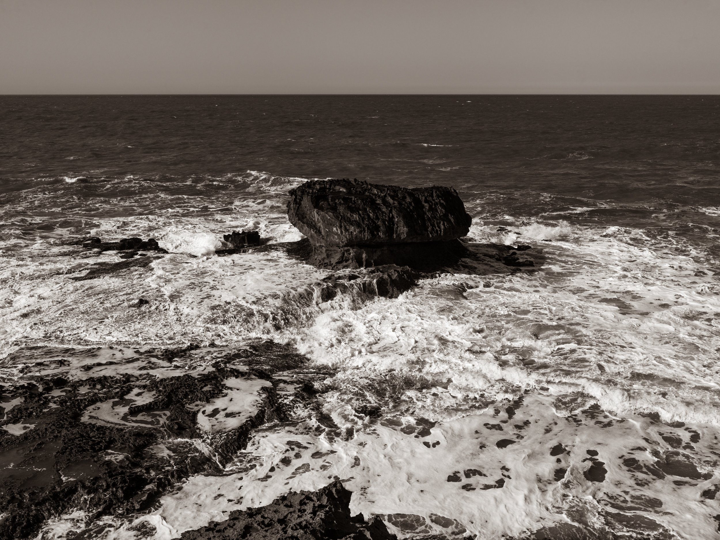 A coastal scene in black and white, featuring waves crashing onto the rocky shoreline and a large rock formation in the water.