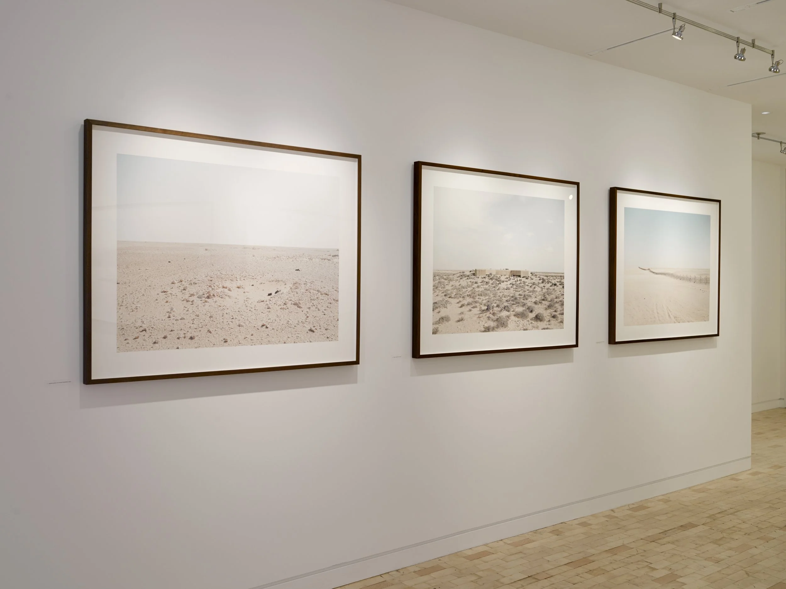 Three framed photographs of desert landscapes with sandy ground and distant structures, displayed on a white gallery wall under track lighting.